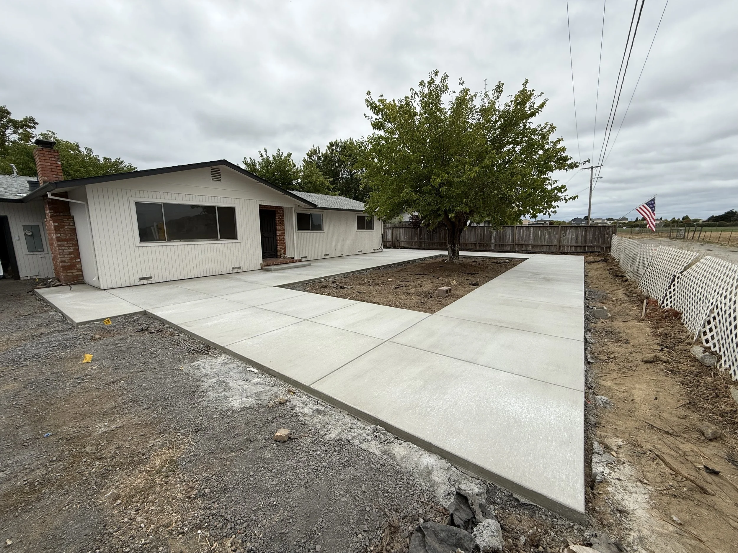 A house with white exterior walls, a brick chimney, and large windows. A newly poured concrete patio in front has a slight step up from the dirt area with some construction debris. A tree with green leaves is nearby, and a wooden fence surrounds the yard. An American flag is flying from a pole on the right side of the image. The sky is overcast with gray clouds.