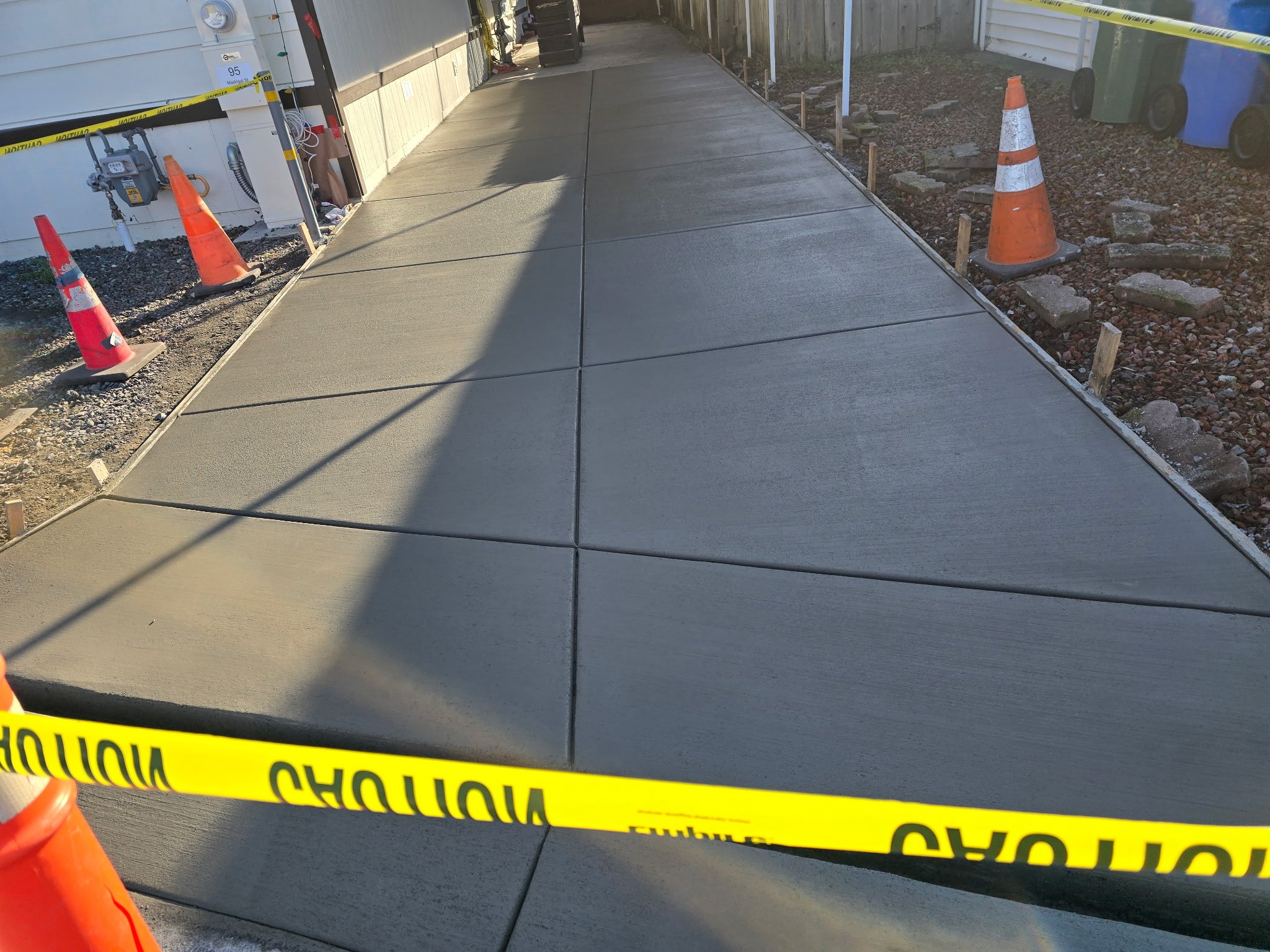Construction area with freshly poured concrete sidewalk cordoned off by yellow caution tape and orange traffic cones.