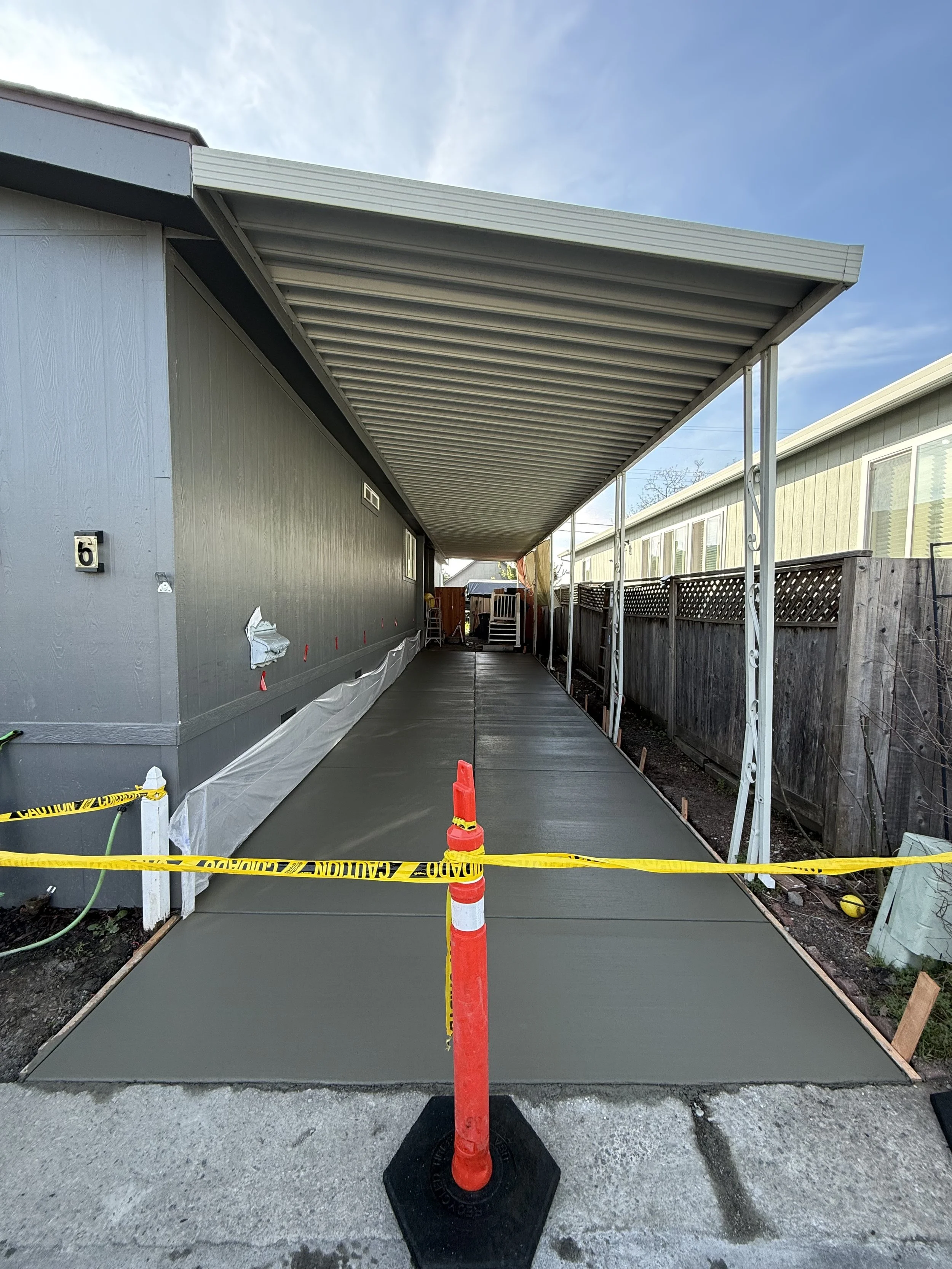 A freshly poured concrete sidewalk under a metal carport with a wooden fence on the right side and a house on the left side. A traffic cone and yellow caution tape are in the foreground, indicating ongoing construction.