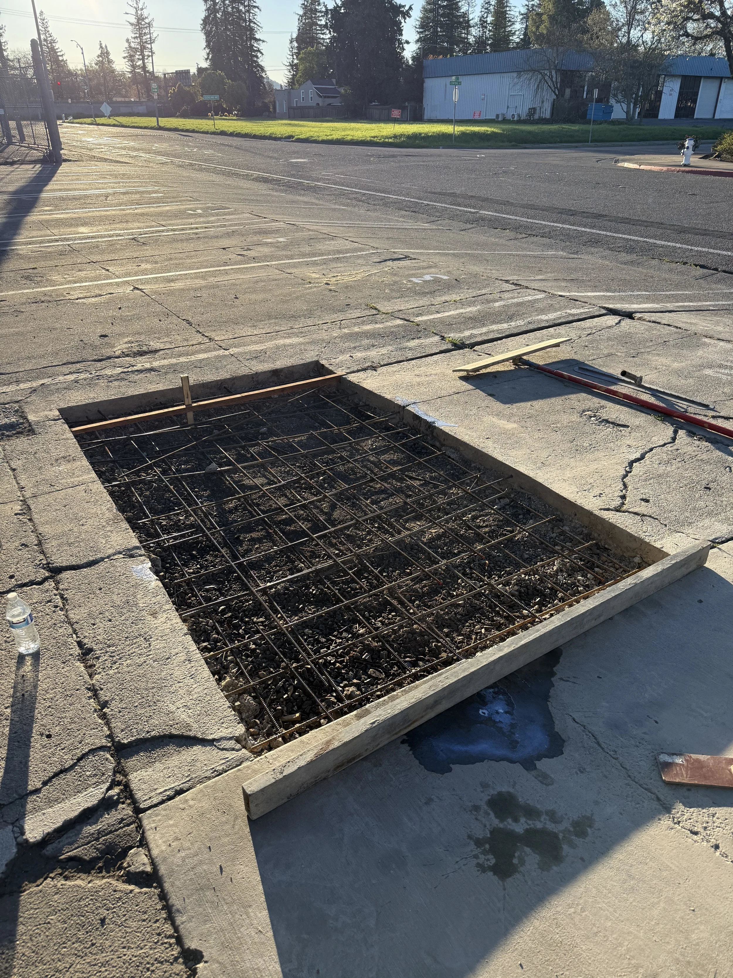 A large sidewalk area under construction with a square excavation marked with wooden framing and metal rebar inside. A water bottle is on the sidewalk to the left, and green grass with trees and buildings are in the background under a clear sky.