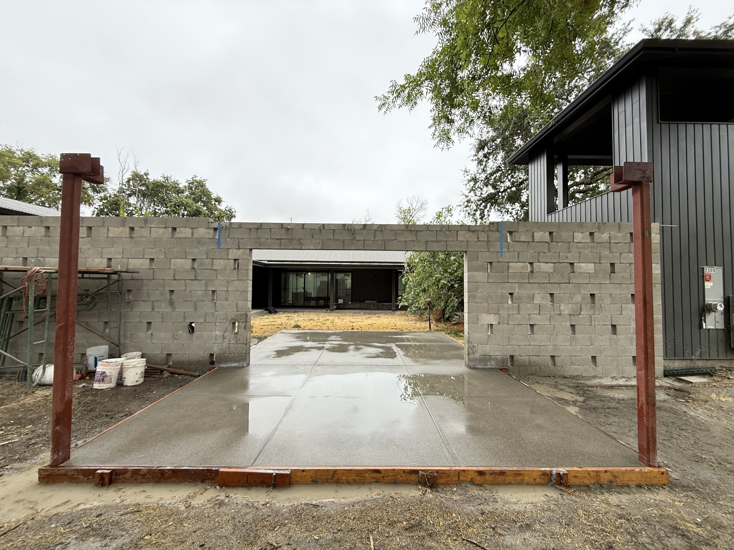 Construction site with a concrete driveway, a cinder block wall, and a black building with a metal siding exterior, surrounded by trees and overcast sky.