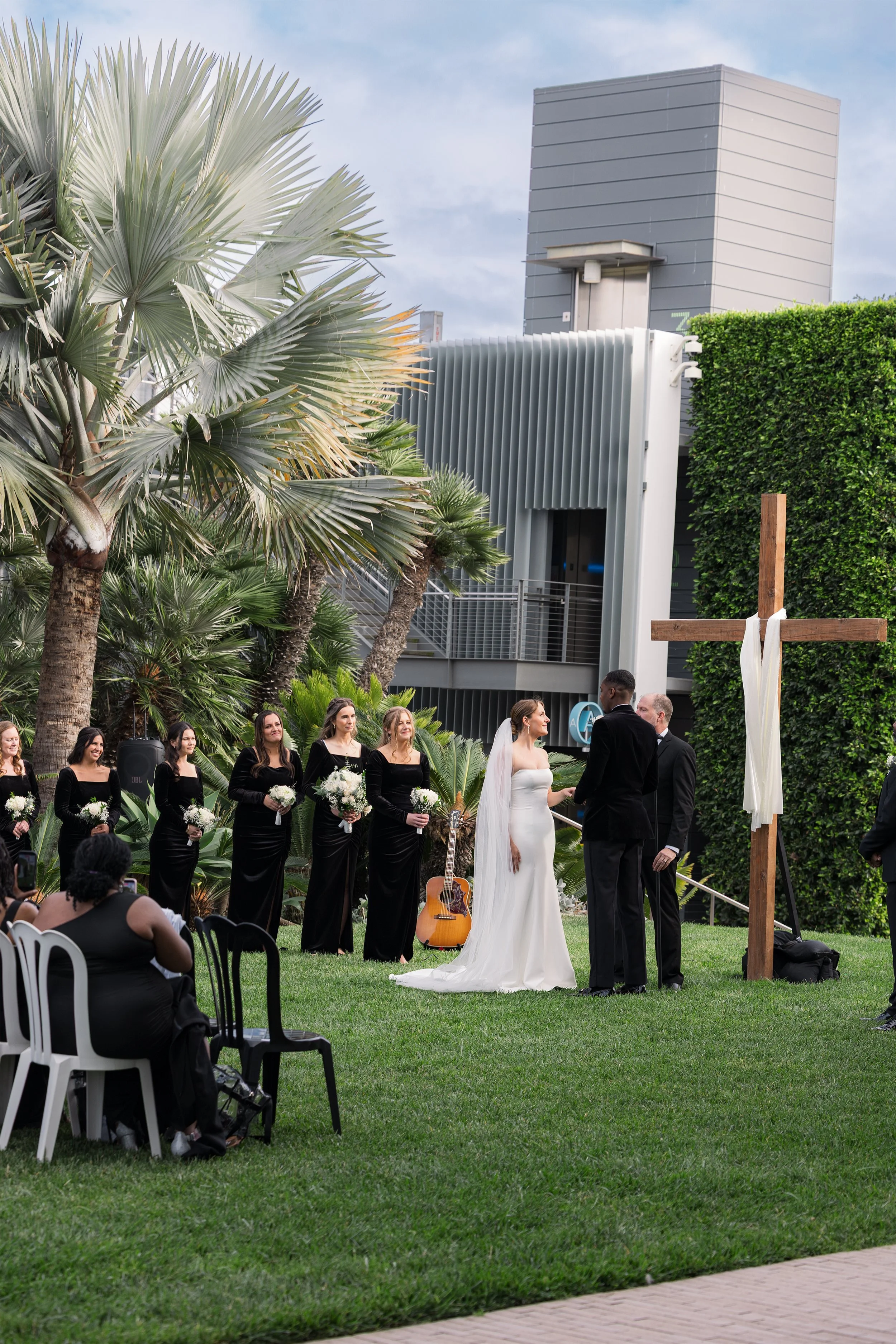 A wedding ceremony taking place outdoors with a bride and groom standing before a cross, surrounded by friends and family. The bride is wearing a white wedding gown and the groom is in a black tuxedo. Bridesmaids in black dresses holding white bouquets and other guests are seated in white and black chairs. There are tall palms, a building, and a hedge in the background.