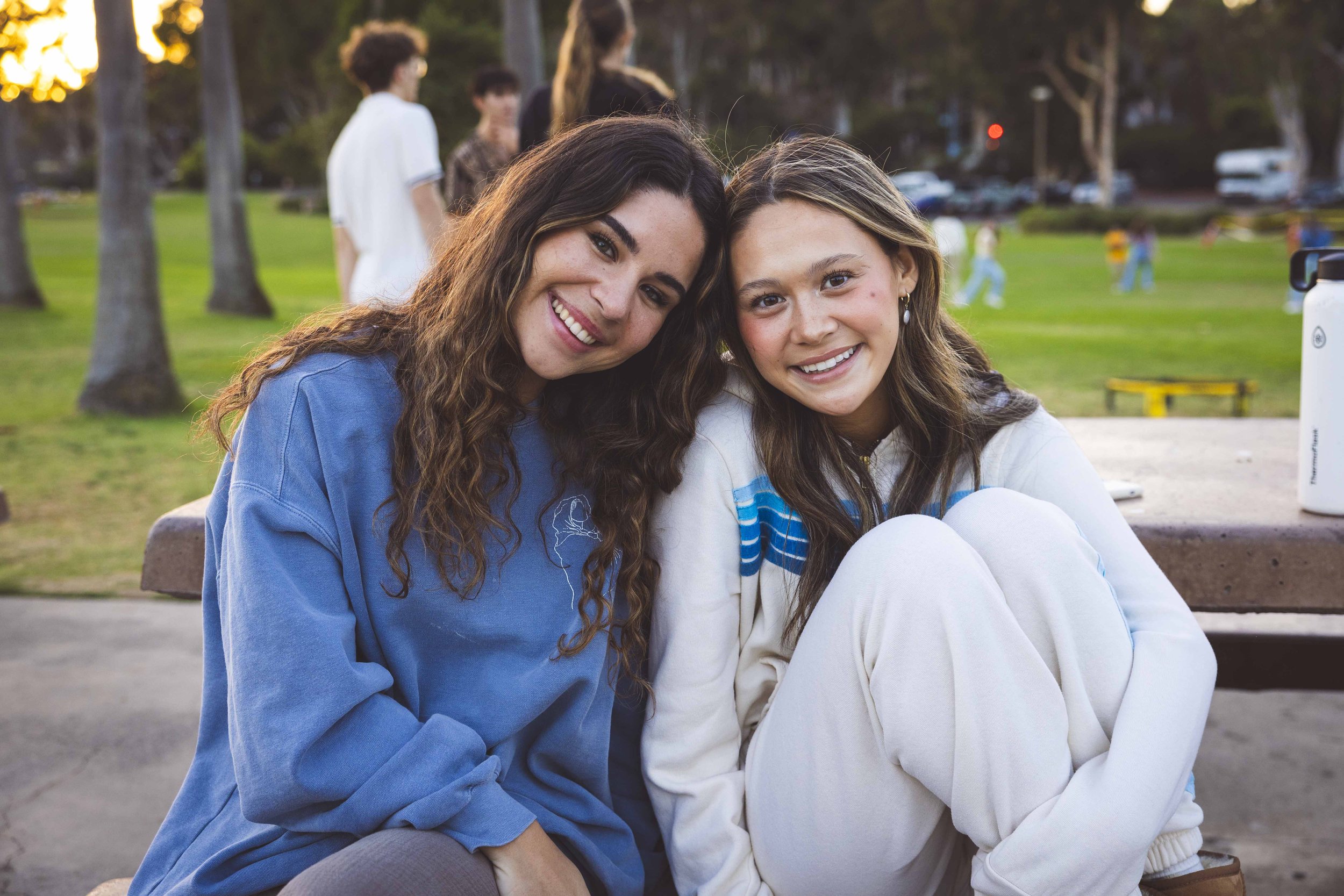 Two young women sitting close together outdoors, smiling at the camera, with trees and other people in the background during sunset.