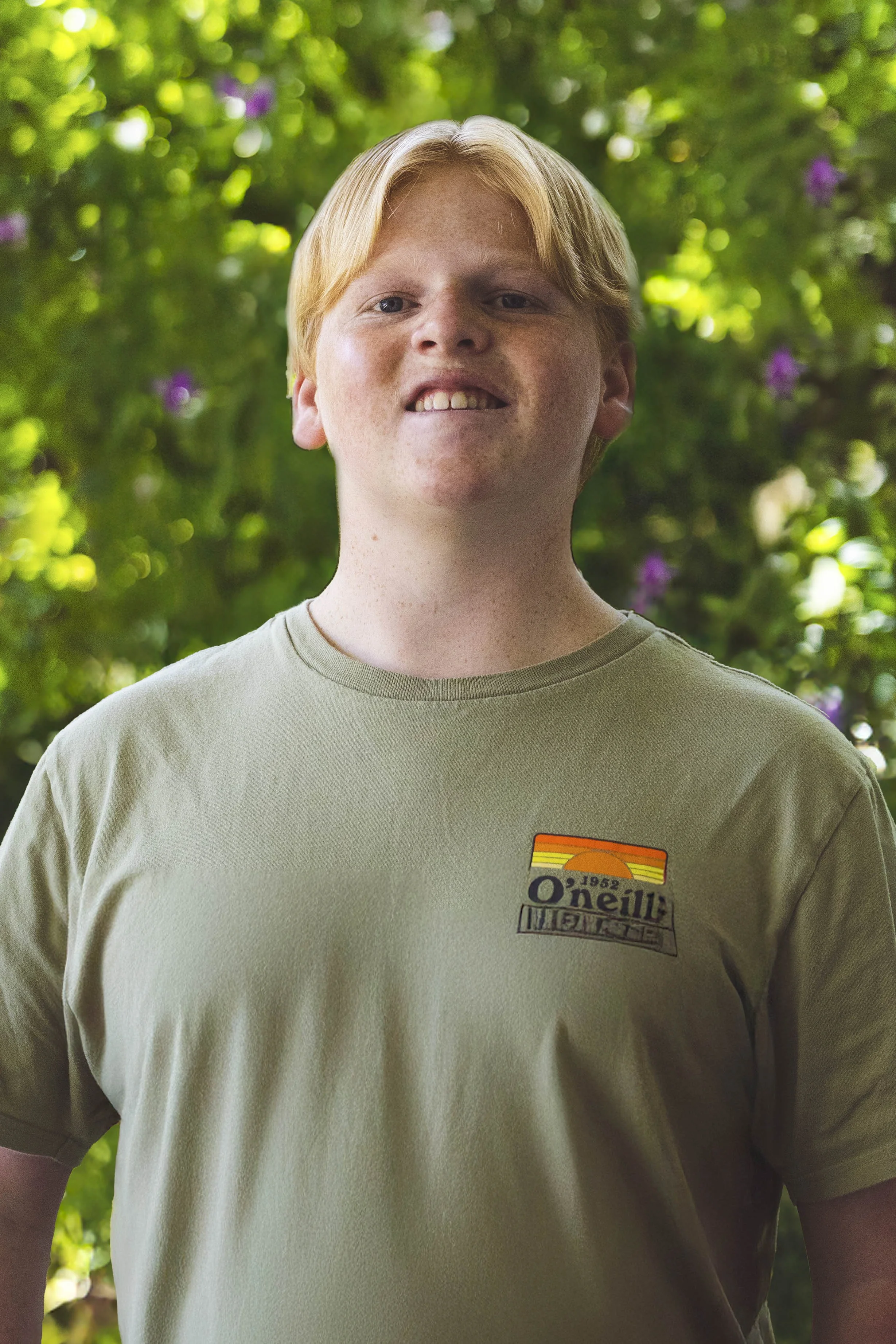 A young man with red hair and freckles wearing a beige t-shirt with a retro O'Neill logo, standing outdoors with green foliage and purple flowers in the background.