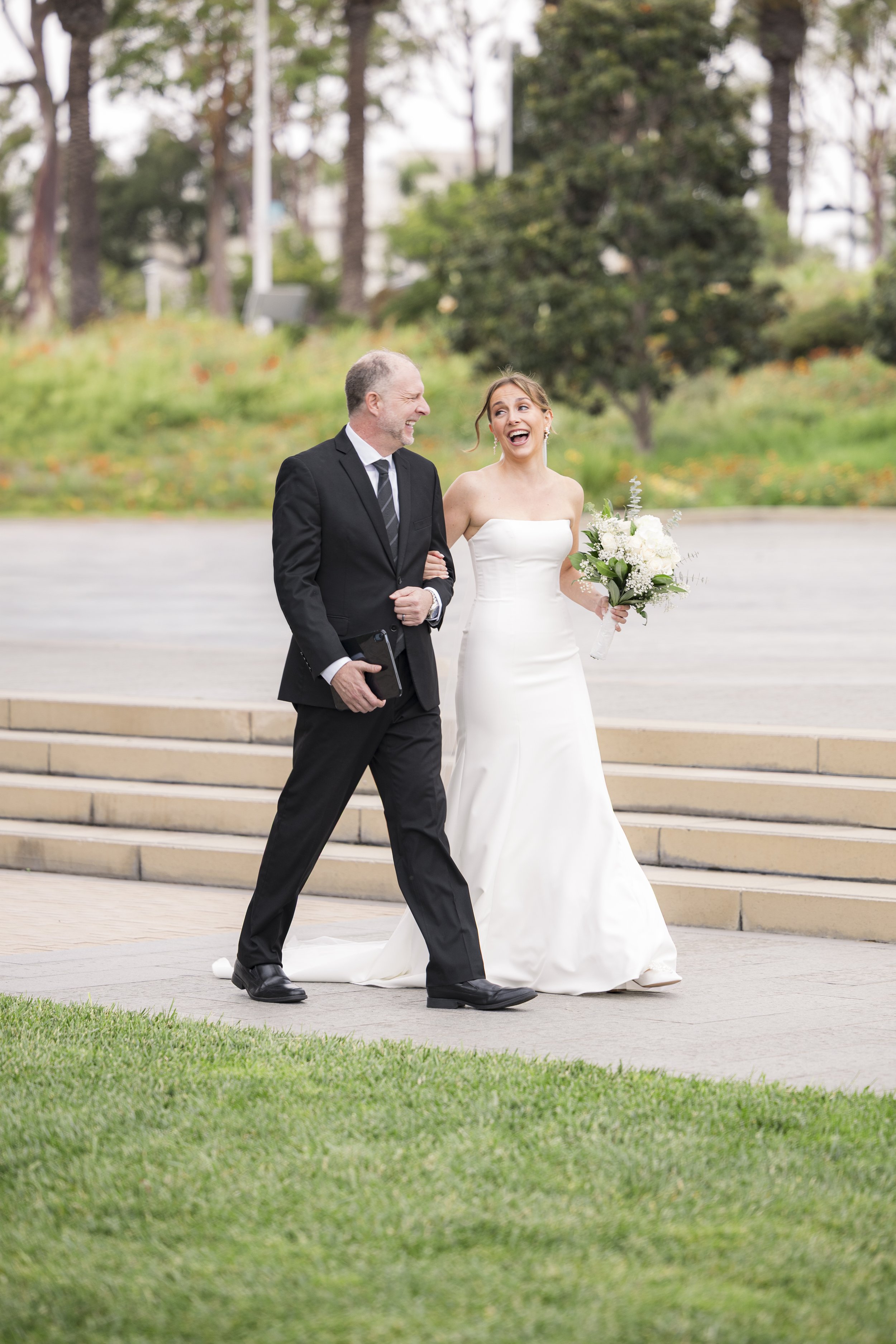 A bride in a strapless white wedding gown holding a bouquet walks arm-in-arm with a groom in a black suit and striped tie on a city street with steps and greenery in the background. They are smiling and laughing.