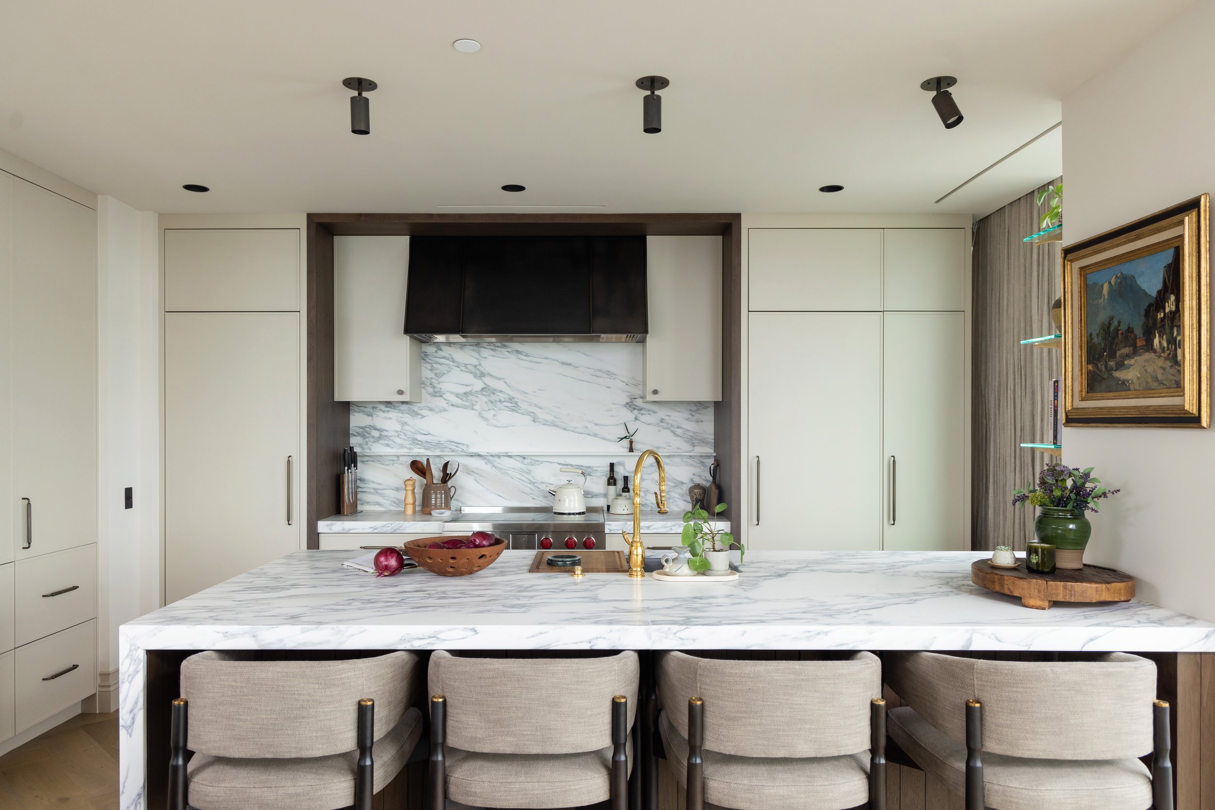 Modern kitchen with white marble island, beige chairs, built-in cabinetry, black top range, marble backsplash, gold faucet, and decorative items including a potted plant and a framed landscape painting.