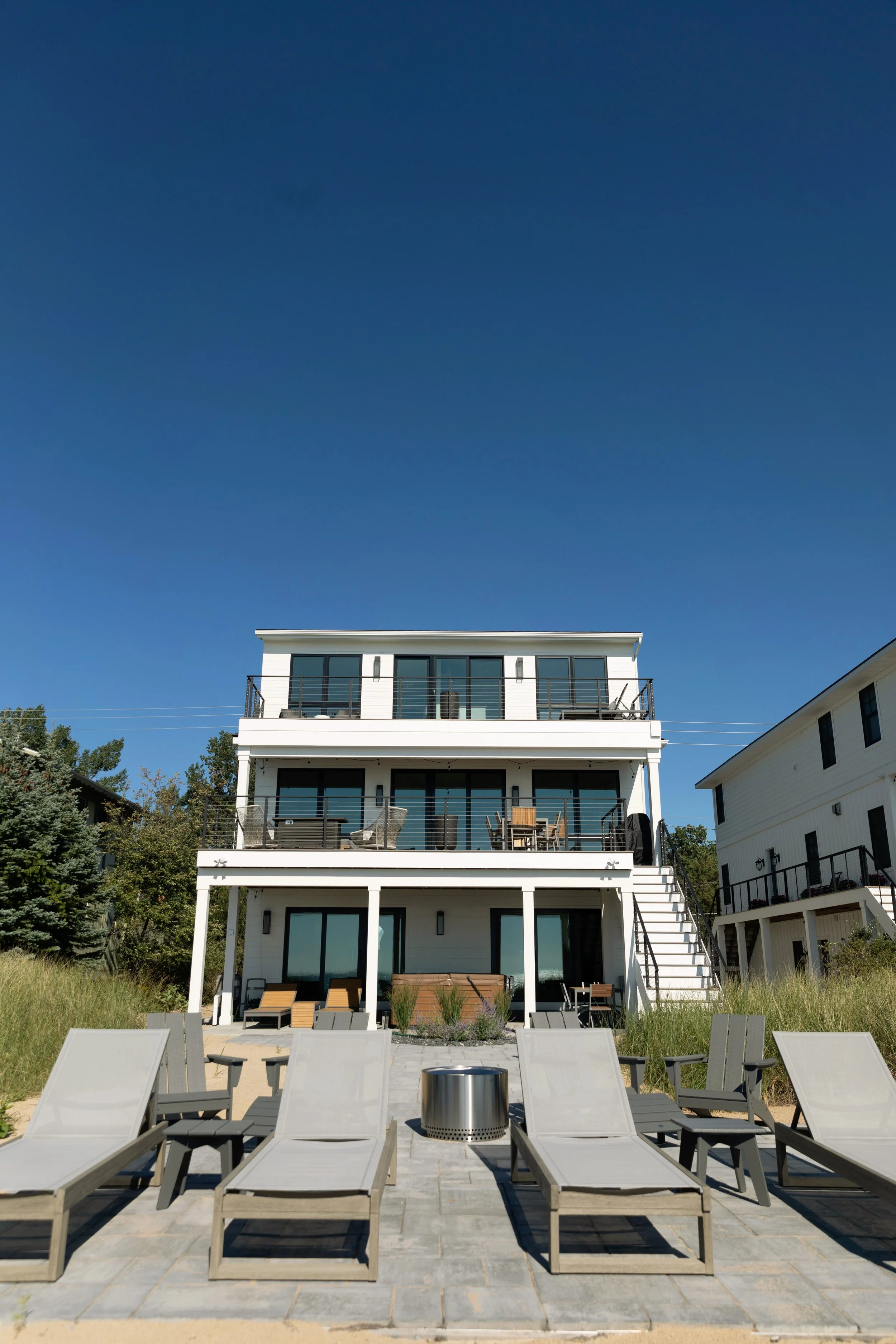 A modern white multi-story house with balconies and large glass doors, set against a clear blue sky. There are lounge chairs and a firepit on a patio area at the backyard, with chairs arranged facing the house.