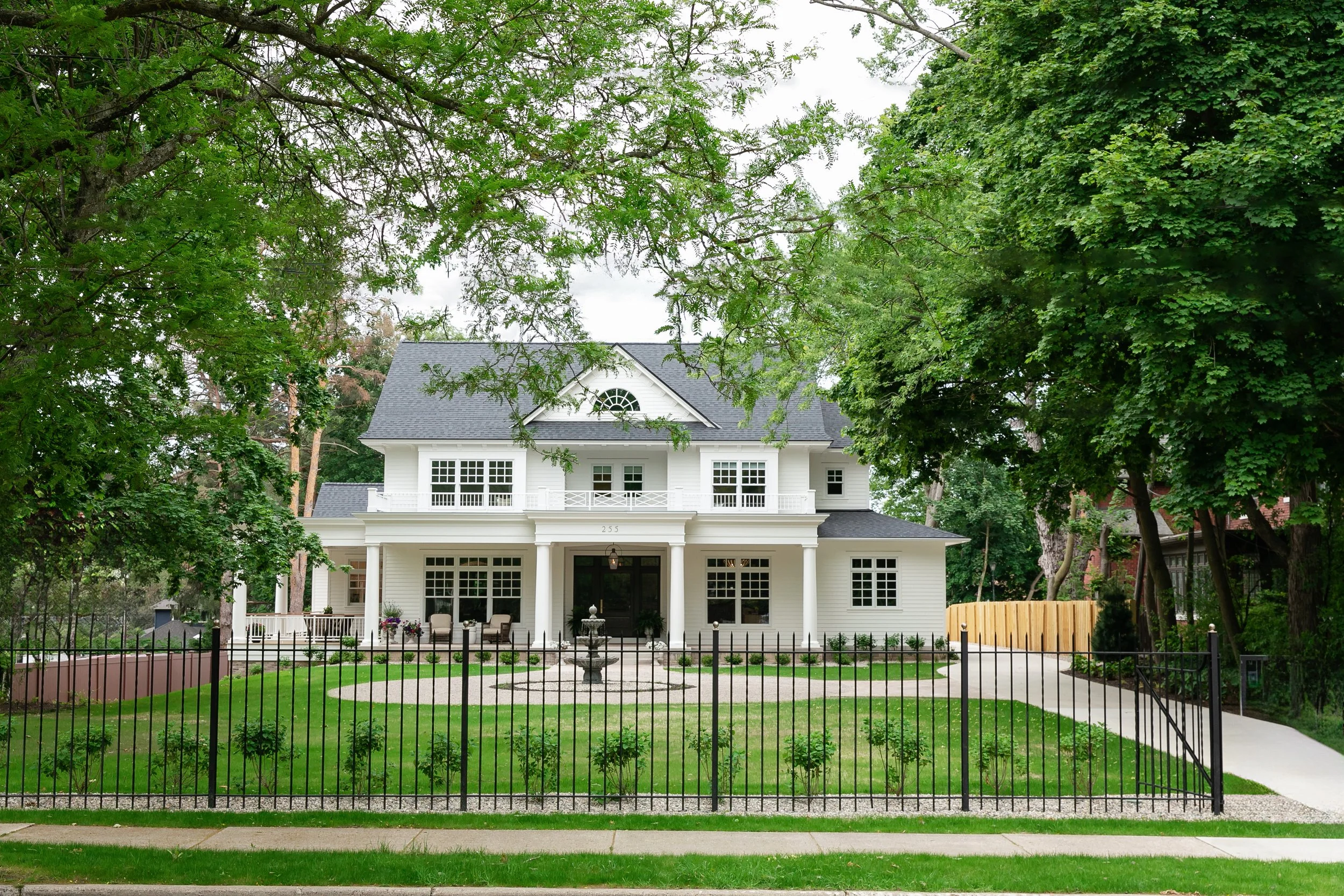 White two-story house with a front porch, surrounded by green trees, a lawn, and a black fence.