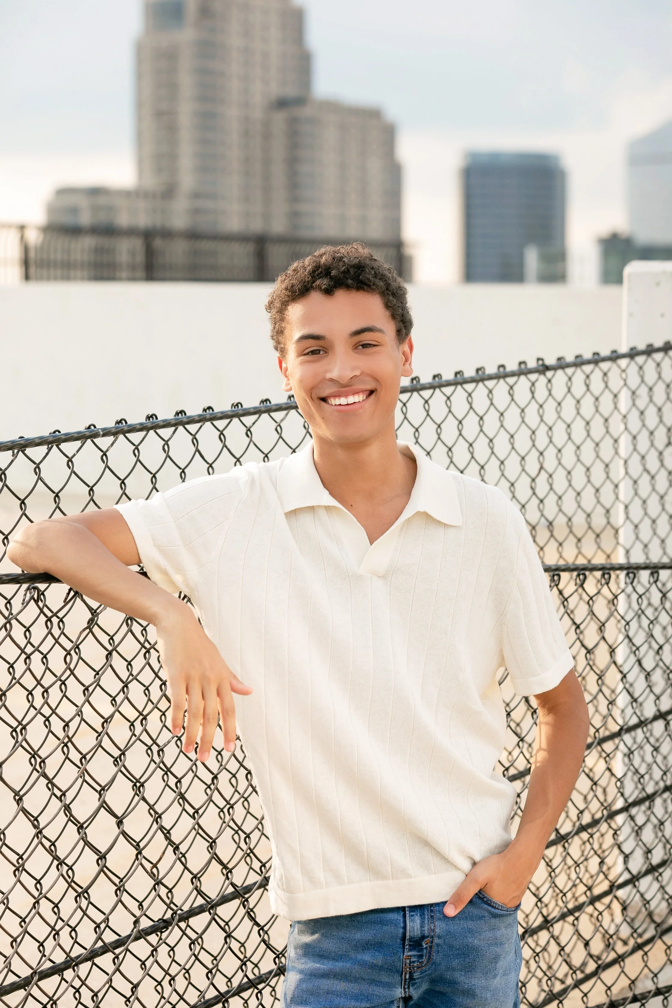 A young man with curly hair smiling, standing outdoors by a chain-link fence with city buildings in the background.