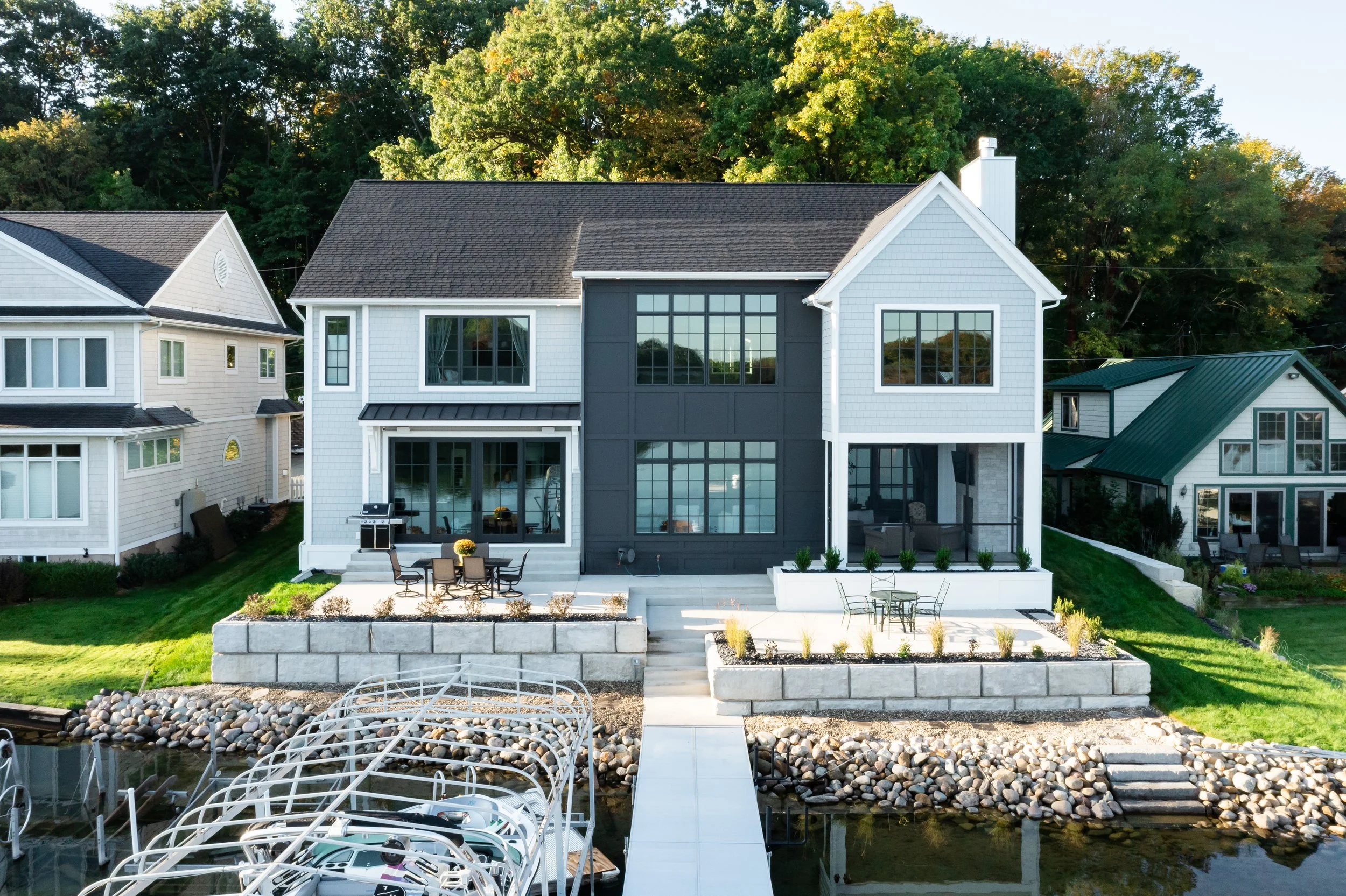 A modern, multi-story house with large windows, a patio with outdoor furniture, and a landscaped yard by a lake with docked boats, surrounded by trees.