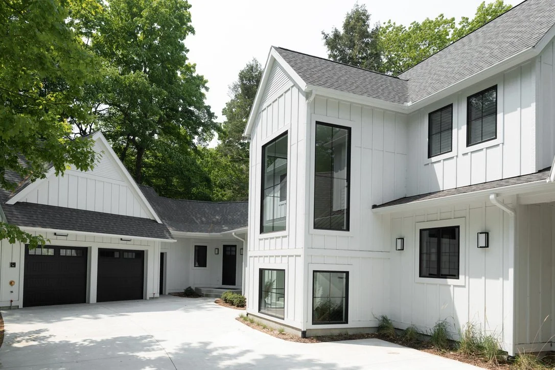 White modern house with black window frames and a three-car garage, surrounded by green trees.
