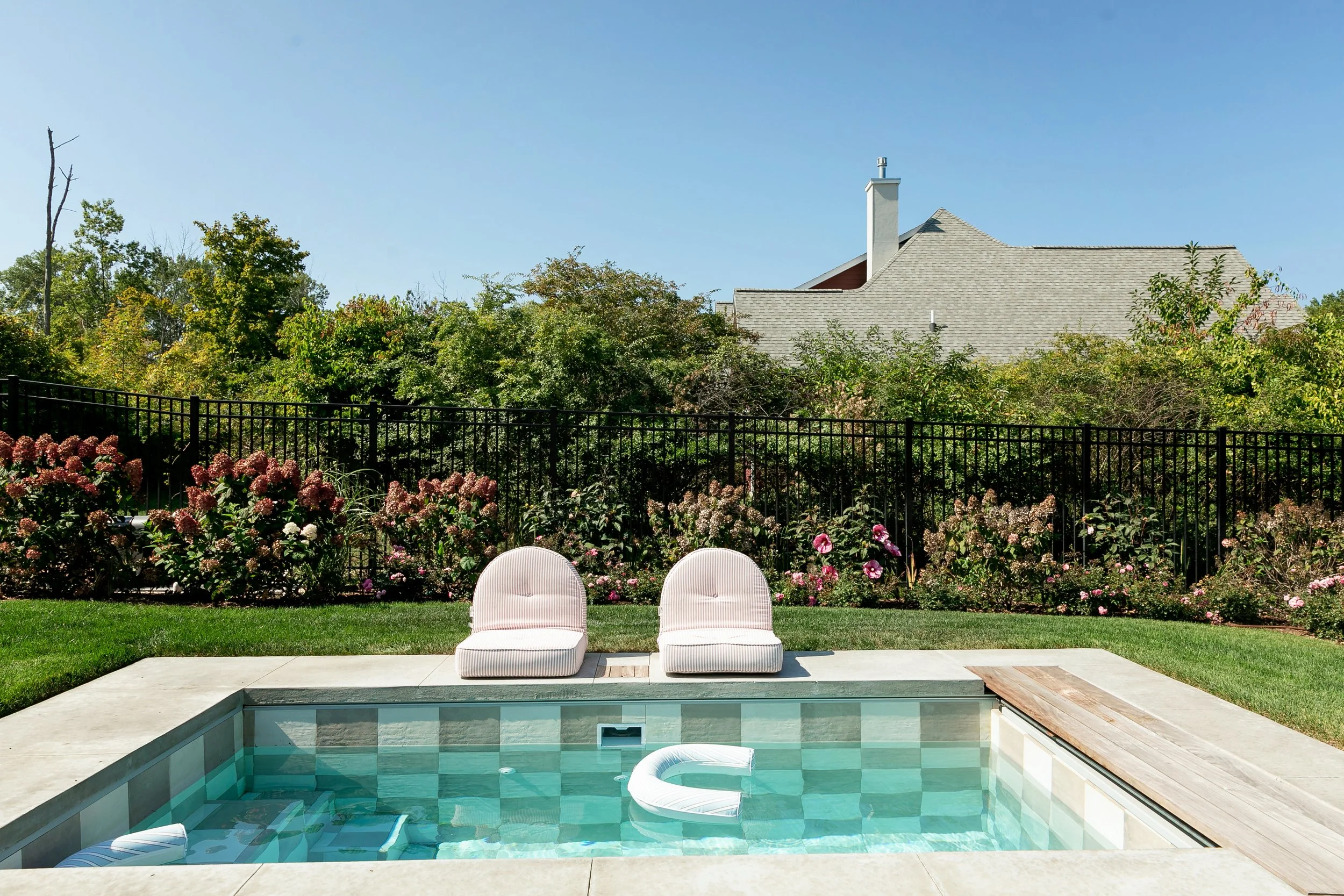 A backyard with a swimming pool, two white lounge chairs, a black metal fence, and a garden with pink and white flowers. In the background, there are green trees and a house with a gray roof against a clear blue sky.