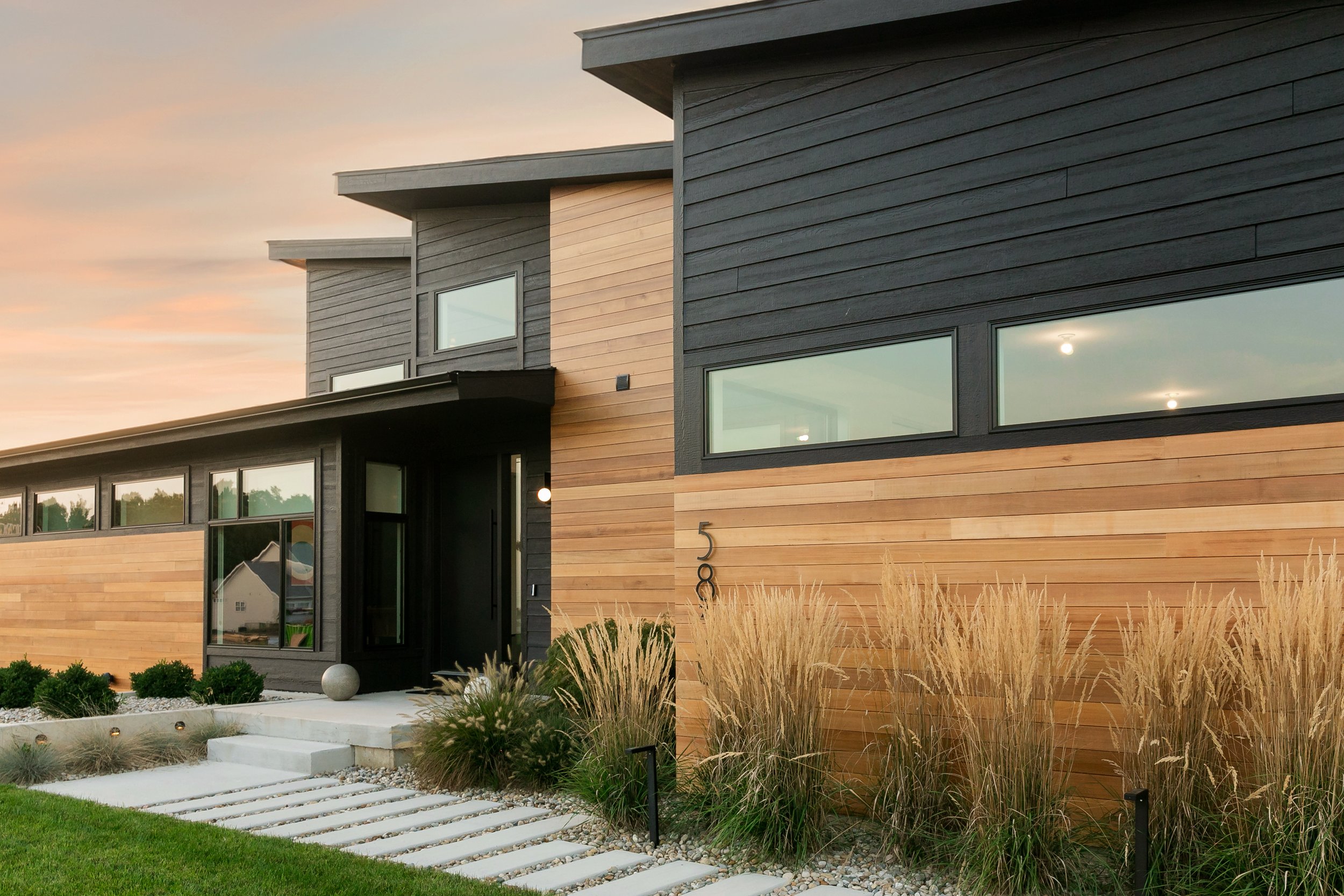 Modern house with wood siding and large horizontal windows, front yard with manicured lawn, stone pathway, and ornamental grasses at sunset.