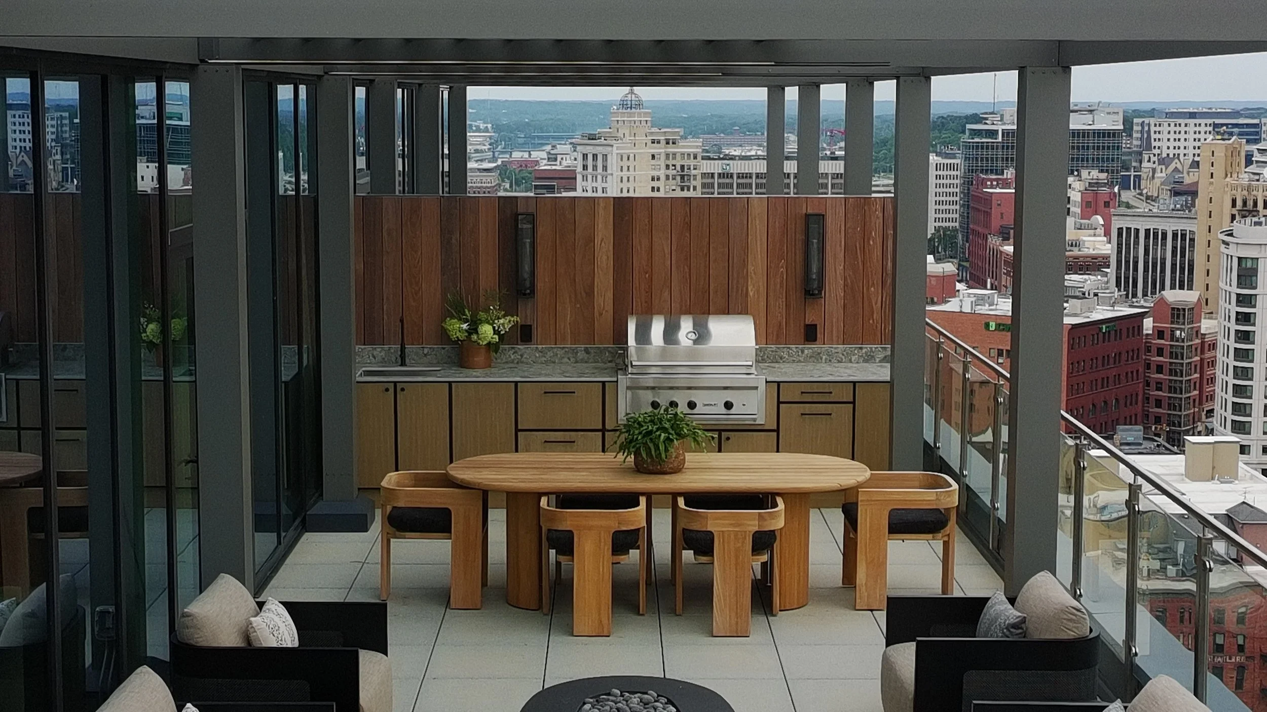 Indoor rooftop patio with a wooden dining table, black chairs, a built-in grill, potted plants, and city skyline view through large glass windows.