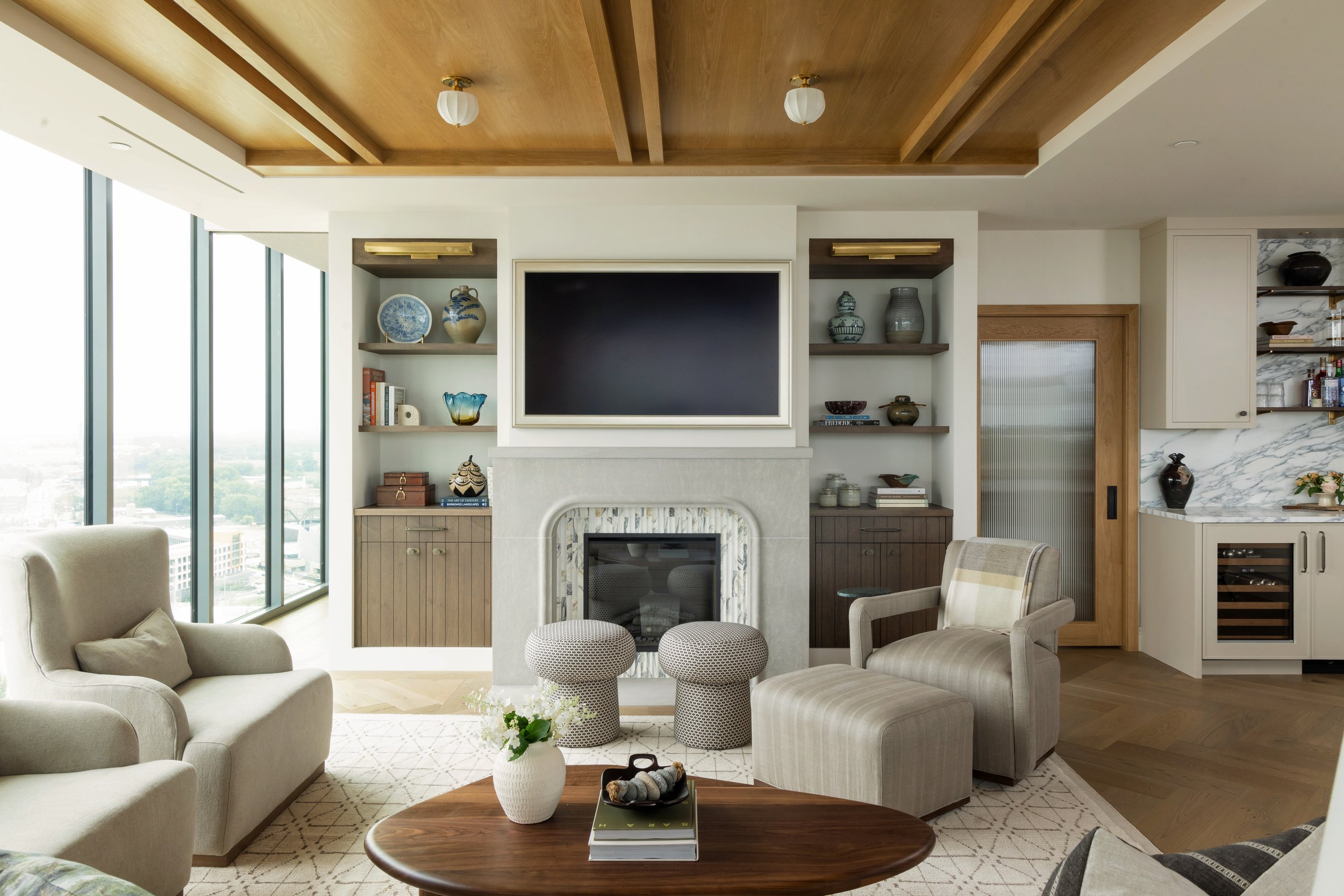 Living room with beige sofas, a wooden coffee table with a white vase and books, a fireplace with a white mantel, built-in shelves with decorative items, and large windows showing a city view.