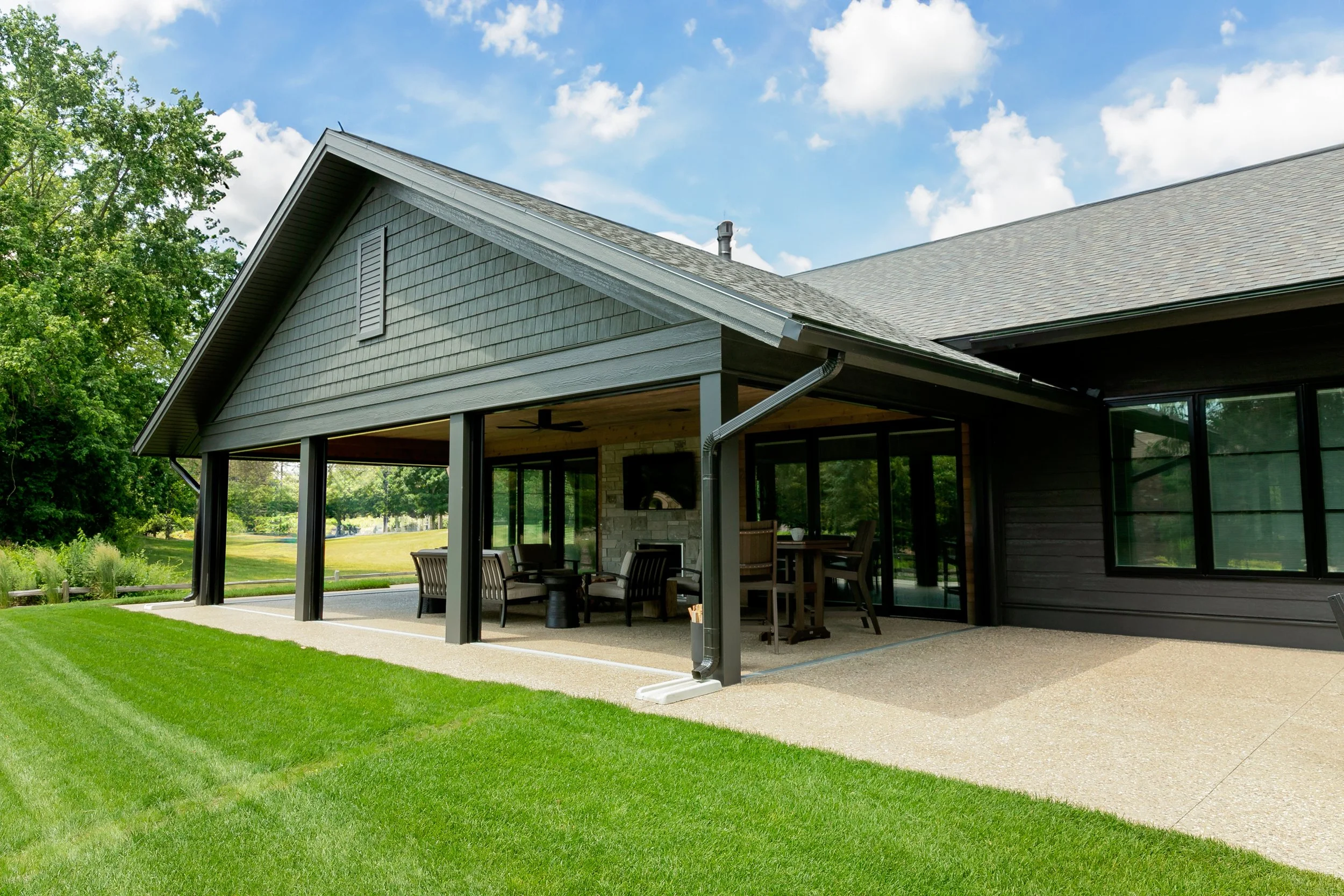 Modern house exterior with a covered patio, outdoor seating, and a well-maintained lawn under a blue sky with clouds.