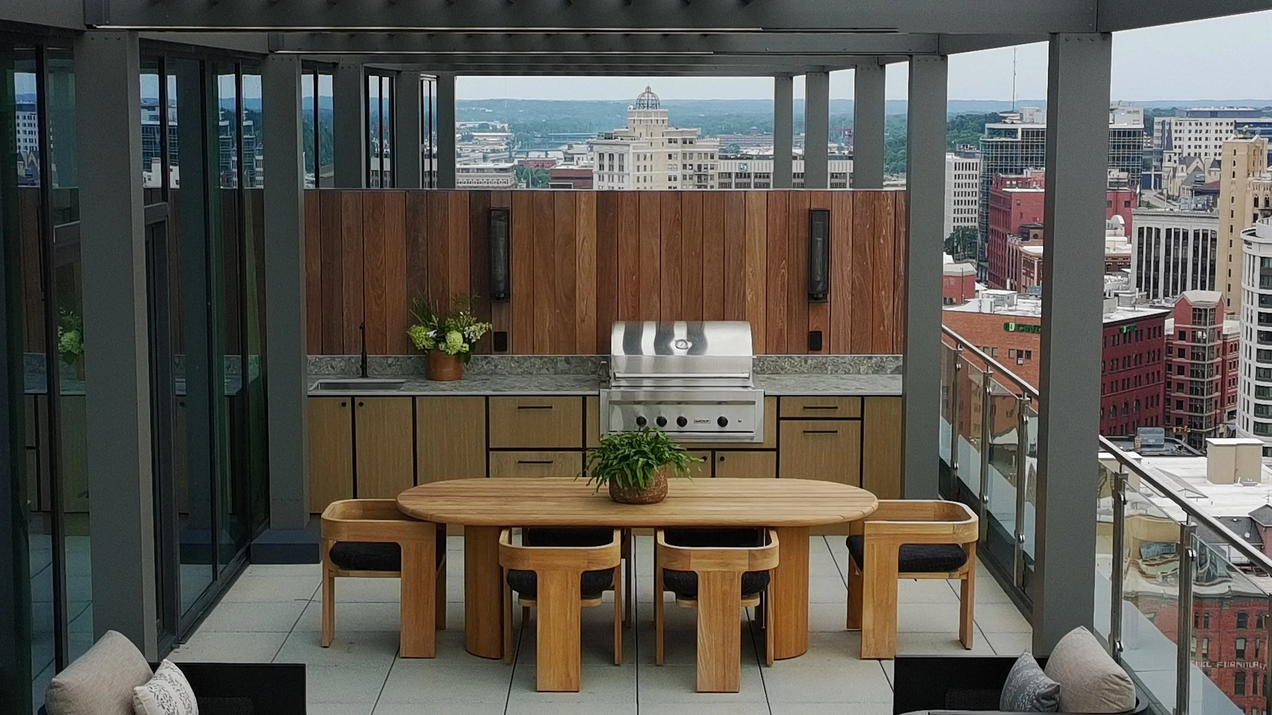 Rooftop balcony with outdoor kitchen, wooden dining table with chairs, potted plants, and city skyline view in the background.