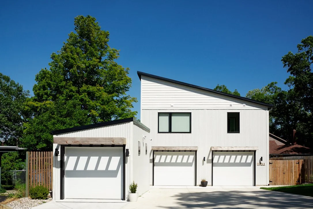Modern white house with three garage doors and a slightly slanted roof, surrounded by green trees and a clear blue sky.