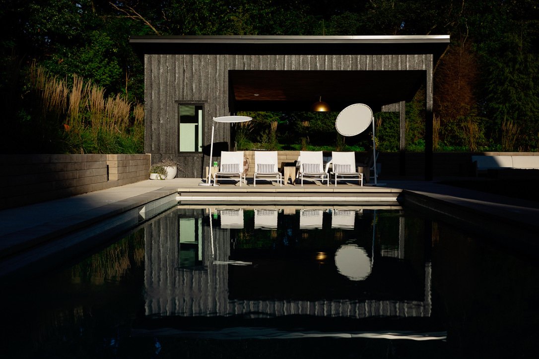 Outdoor patio with six white chairs, two round patio umbrellas, and a large reflector, near a swimming pool, with a dark wooden house and greenery in the background.