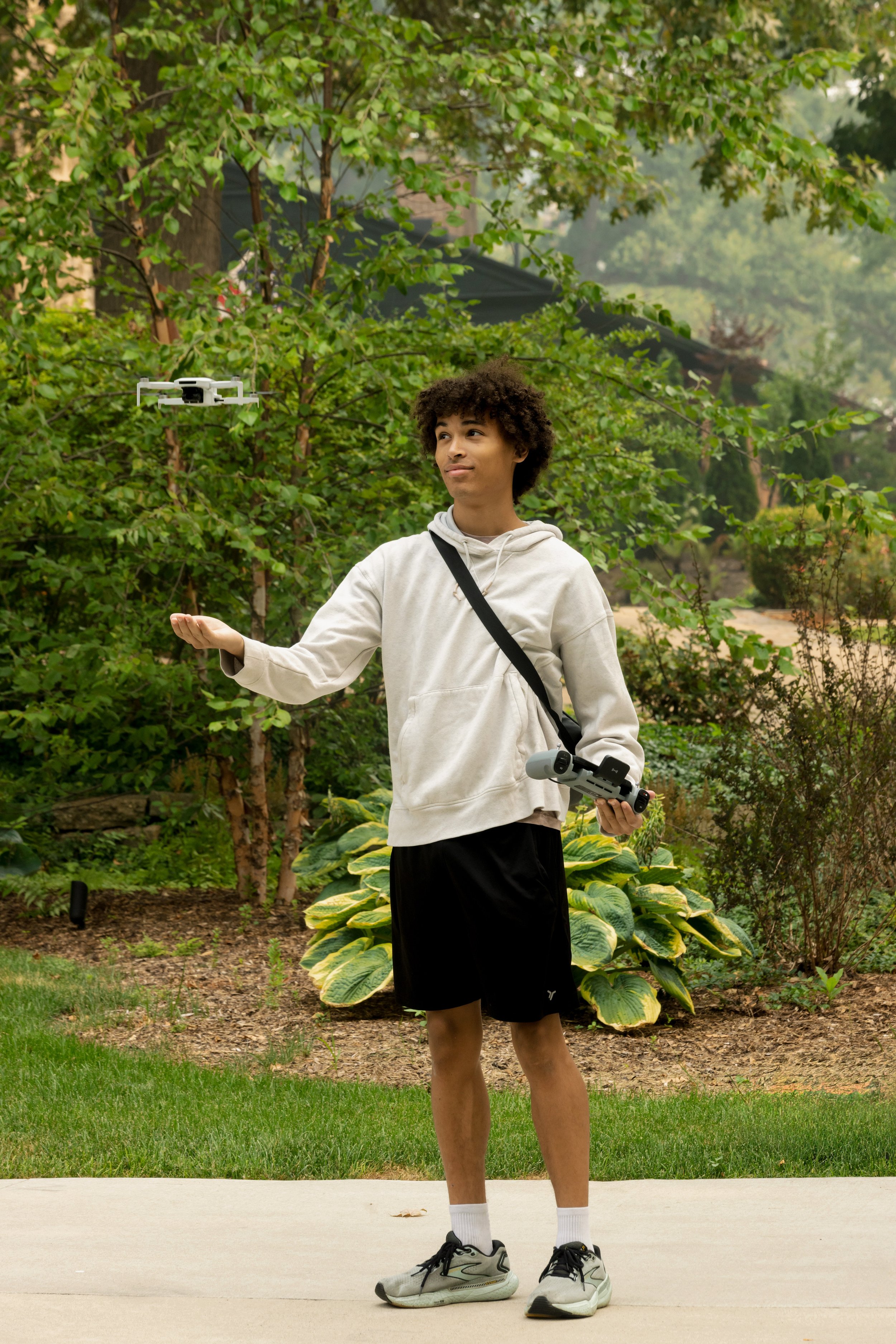 A young man stands outdoors on a sidewalk holding a drone remote control in one hand and a drone in the other. He is wearing a beige hoodie, black shorts, white socks, and sneakers. Behind him are lush green trees and plants.