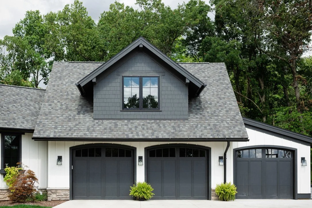 A modern house with gray garage doors, white walls, black window frames, and a dark gray roof, surrounded by green trees and shrubs.