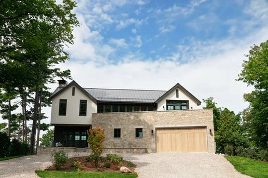 Modern two-story house with a combination of stone and white siding exterior, a garage, surrounded by greenery and trees, under a partly cloudy sky.