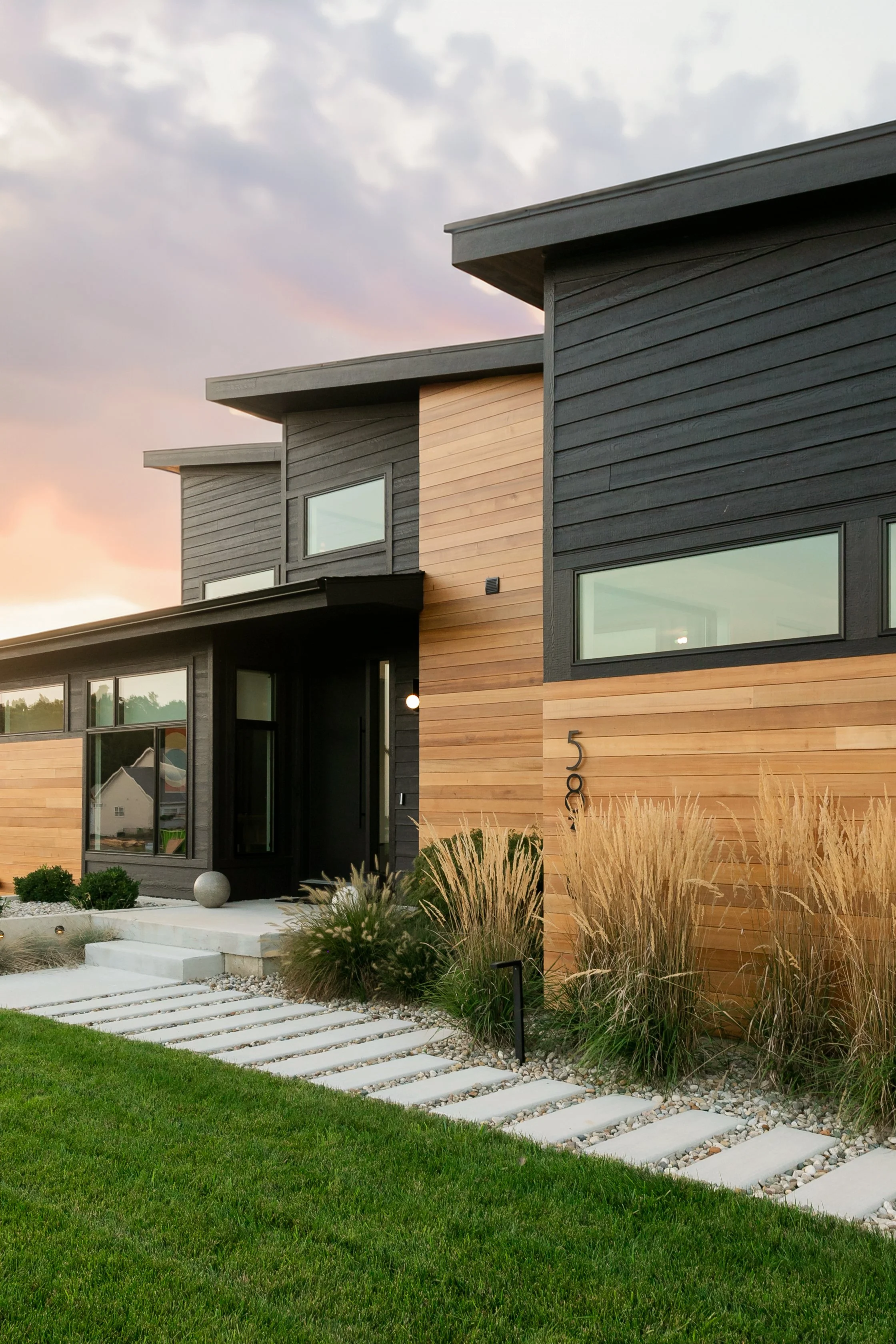 Modern house with black and natural wood siding, large windows, a concrete walkway with steps, and landscaping with grasses and shrubs, under a partly cloudy sky at sunset.