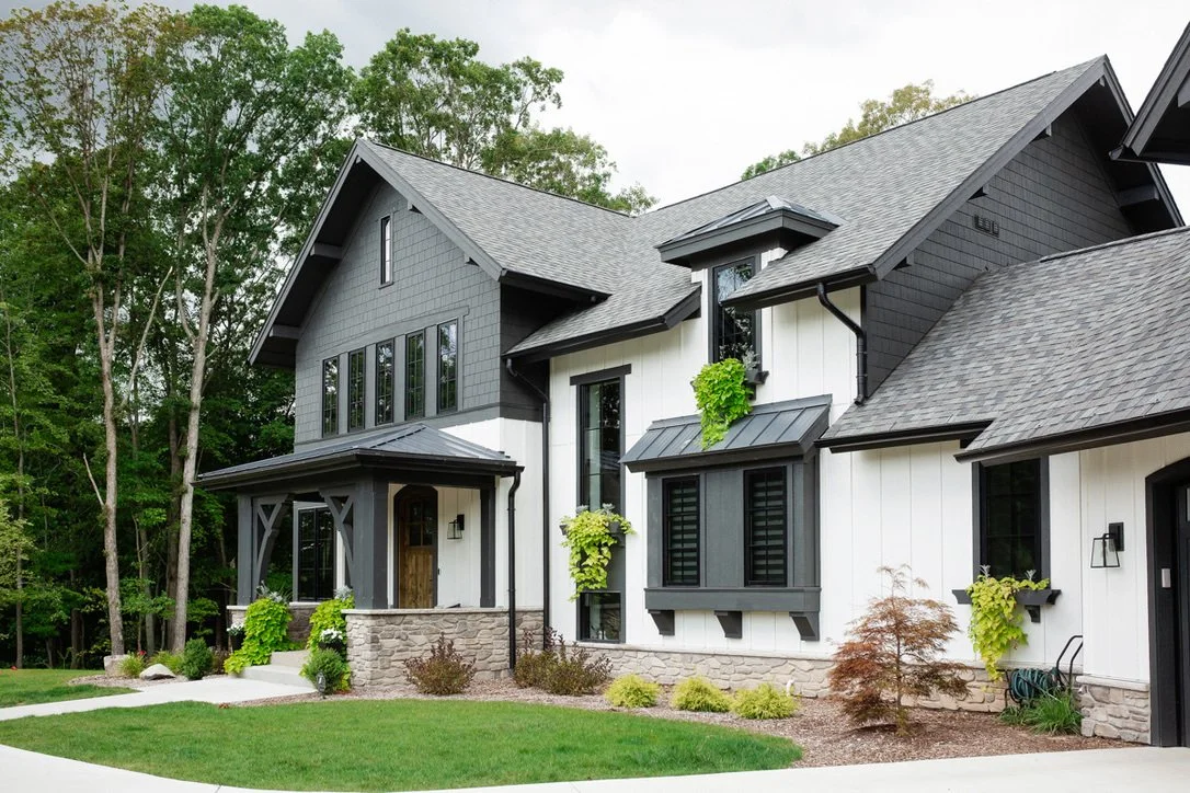 Modern two-story house with black and white exterior, stone accents, and lush landscaping, set in a wooded area.