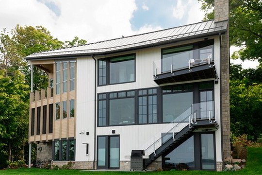 Modern three-story house with large windows, balconies, and a metal roof, surrounded by trees and greenery.
