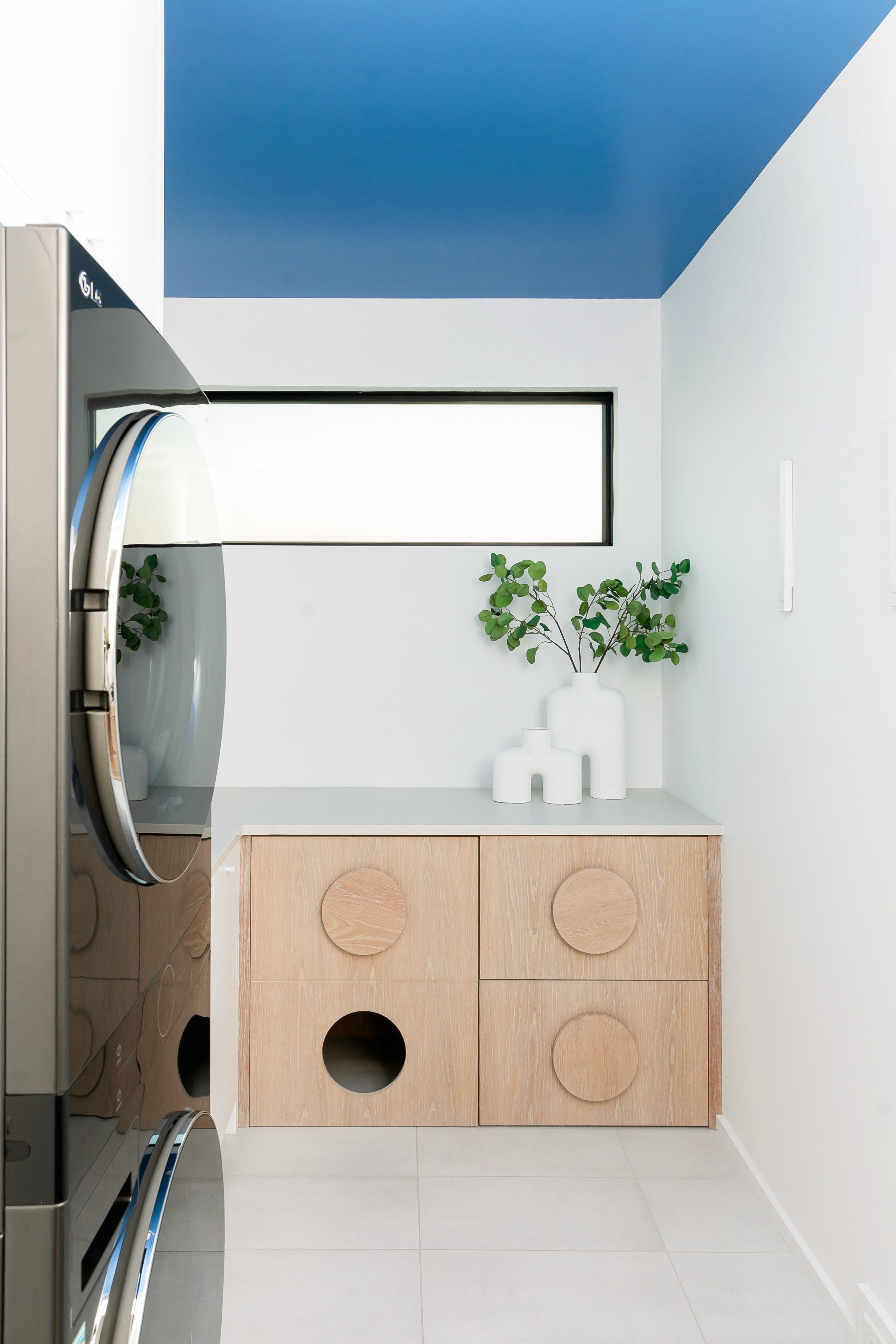 A modern laundry room with a wooden cabinet featuring circular cut-outs and a white vase with green leafy branches on top, a horizontal window, and a blue ceiling.