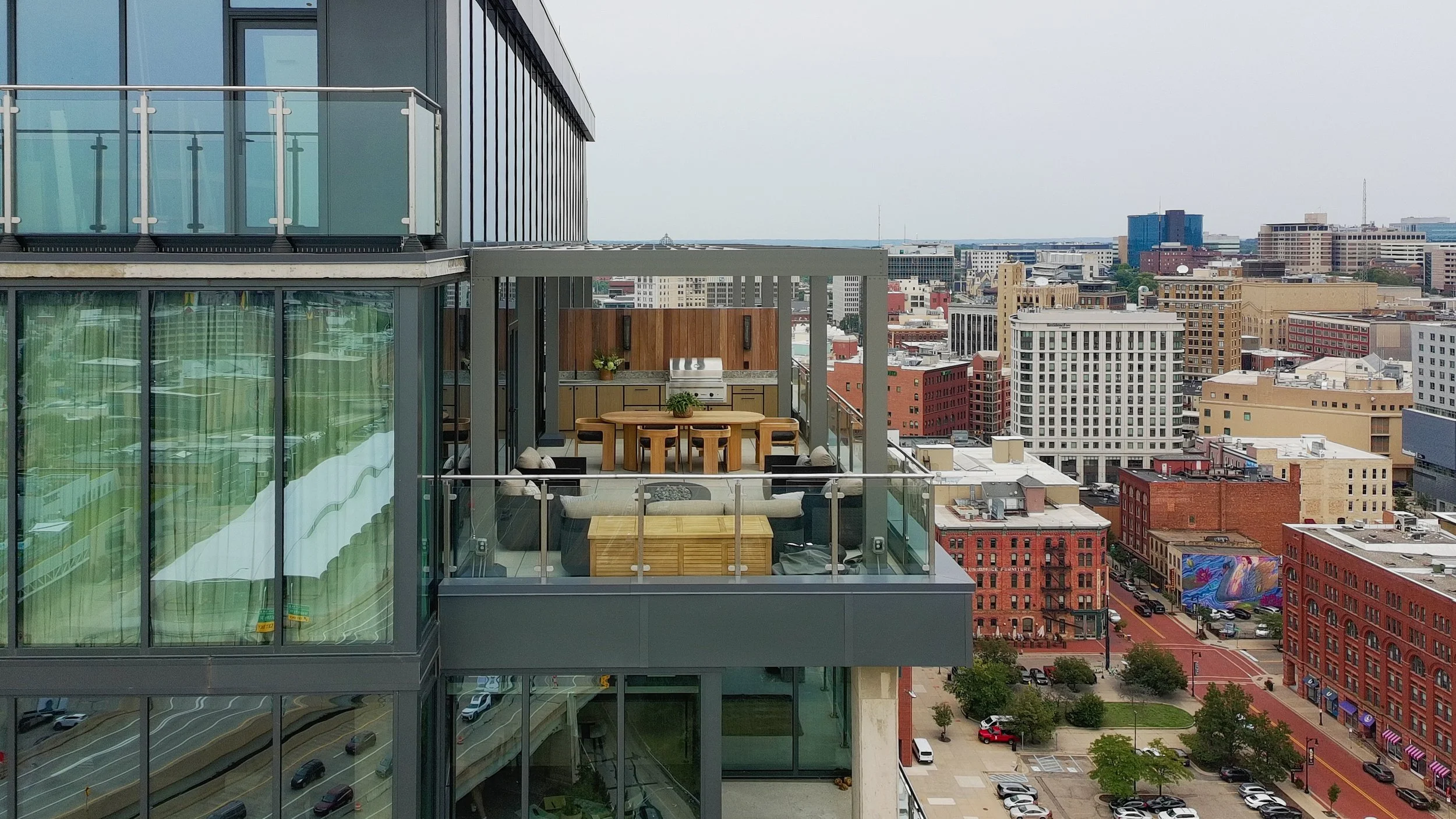 Rooftop balcony with outdoor furniture, a grill, and city skyline view including various buildings and streets.