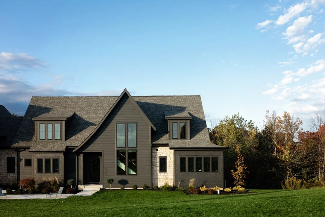 A two-story house with gray siding, multiple windows, and a steep roof, set in a green yard with trees in the background.
