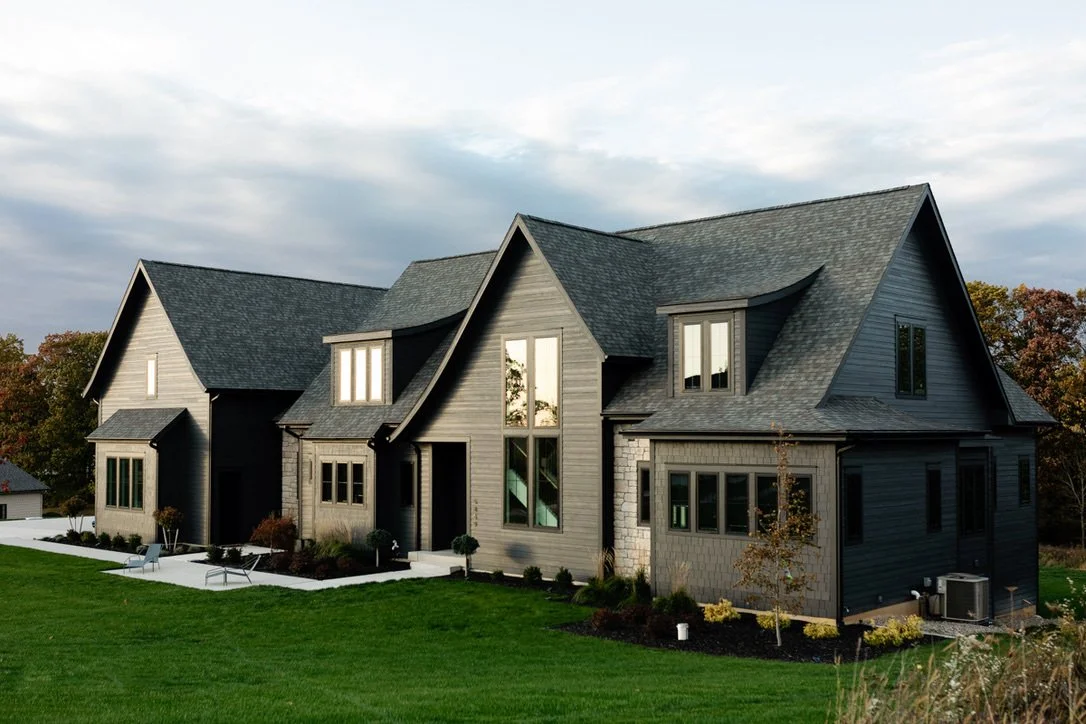 A modern black house with multiple gabled roofs and large windows, surrounded by a well-maintained lawn and some small trees.