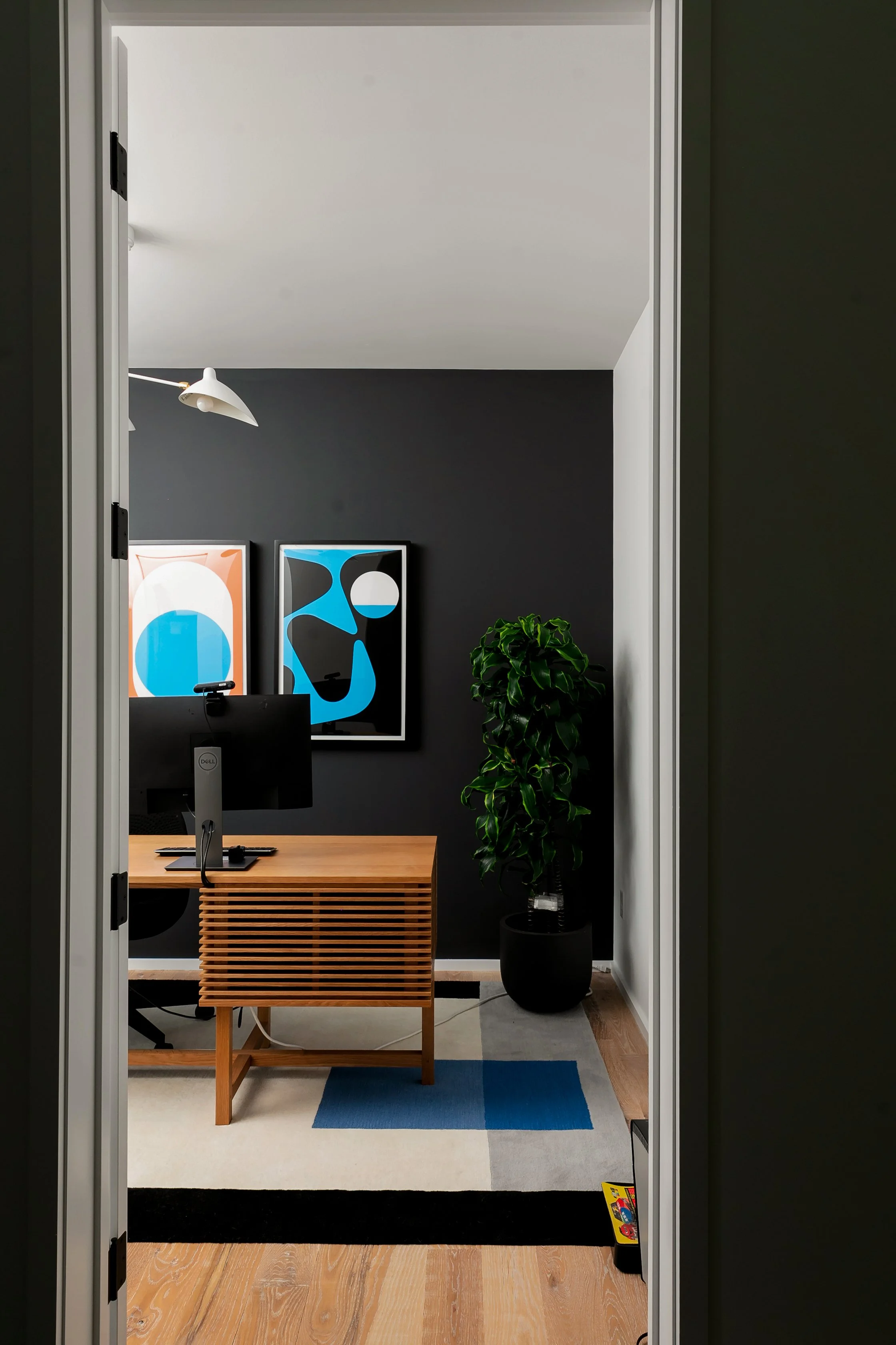 View of an office with a wooden desk, a computer monitor, two colorful abstract art prints on a dark wall, a green leafy plant, a floor lamp, a geometric rug, and wooden flooring.