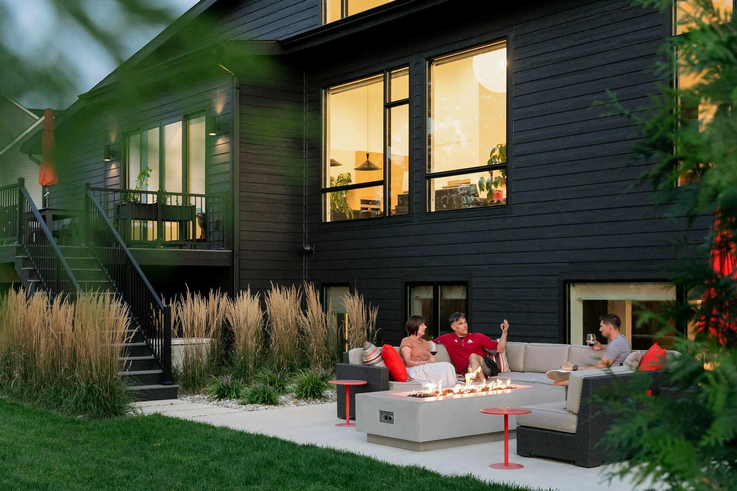 A group of people sitting on outdoor furniture around a fire pit in a backyard at dusk, with a modern black house in the background and city lights in the distance.