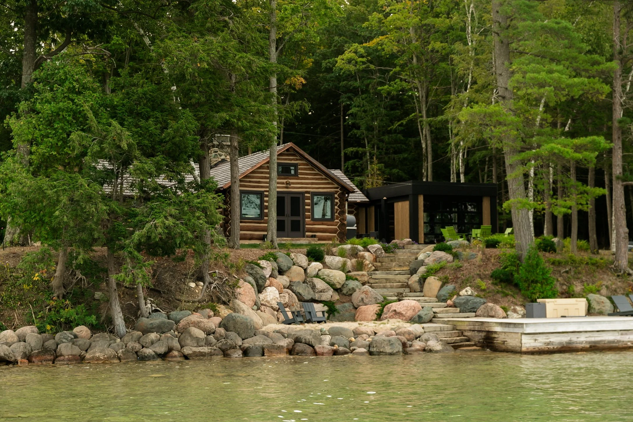 A lakeside scene with a cabin built of logs, surrounded by tall green trees, with a stone pathway leading up to it and chairs set outside; the water in the foreground is calm.