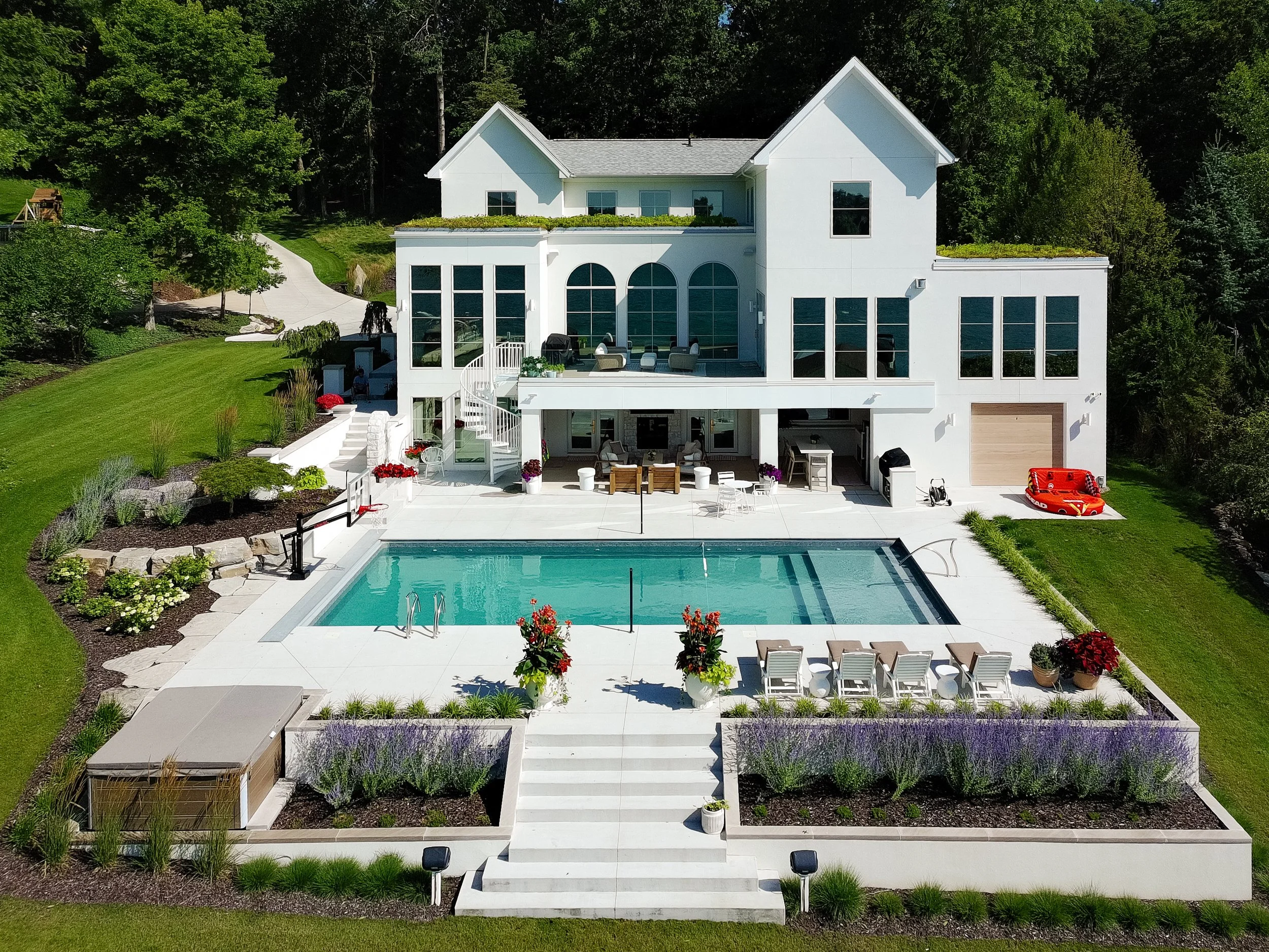 A large modern white house with multiple levels and large windows, surrounded by a lush green lawn and landscaped garden, with an outdoor pool and patio area in the backyard.