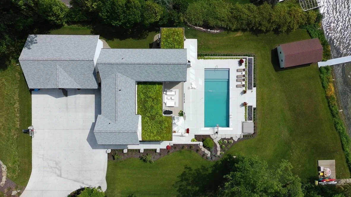Aerial view of a house with a gray roof, a swimming pool, a lawn, and surrounding trees.
