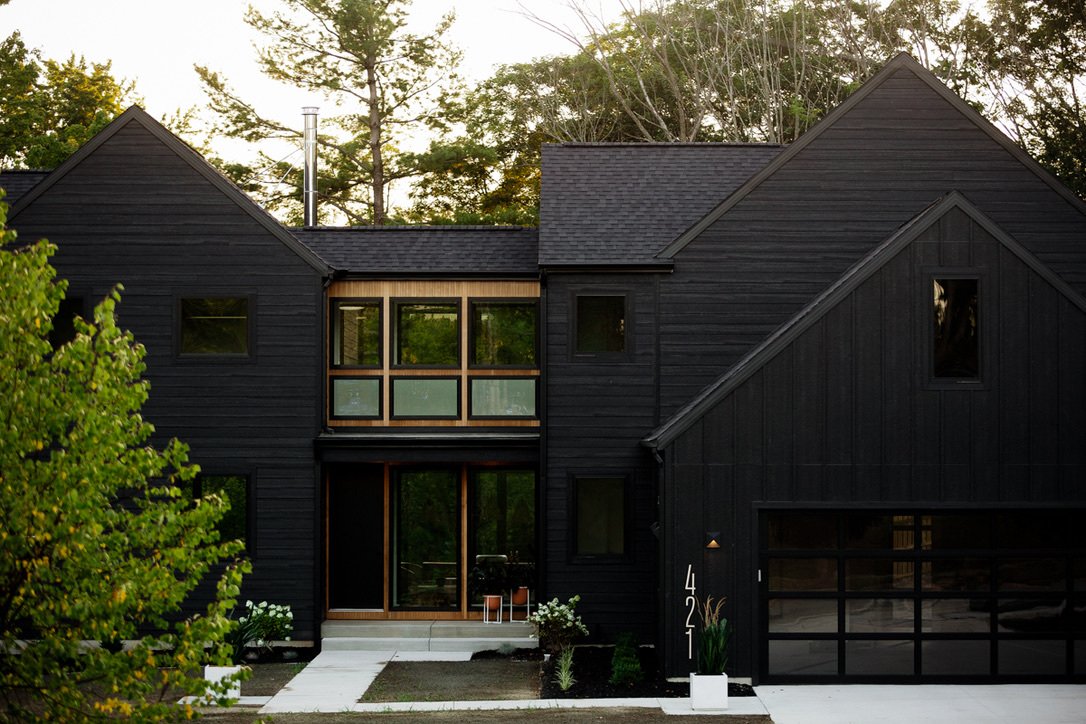 Modern black house with large windows and a greenhouse extension, surrounded by trees and green foliage.
