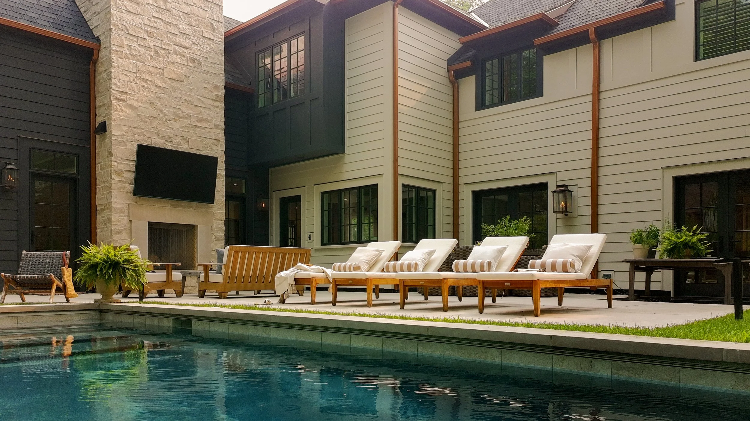 A backyard with a swimming pool, outdoor lounge chairs with striped cushions, potted plants, a stone fireplace, and a modern house with large windows and siding, during daytime.
