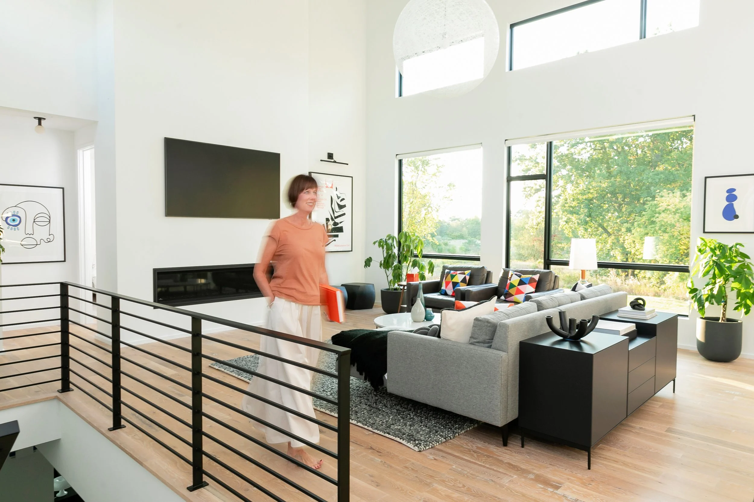 A woman standing in a modern, bright living room with large windows, grey sofas with colorful cushions, and contemporary artwork on the walls.