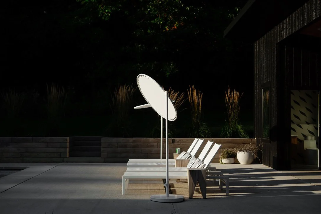Outdoor patio with four white lounge chairs, a large white umbrella, potted plants, and a dark building wall at night.