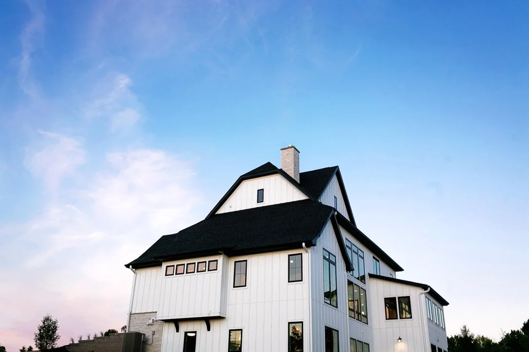 A large, modern white house with black roof shingles, multiple windows, and a tall chimney against a clear blue sky.