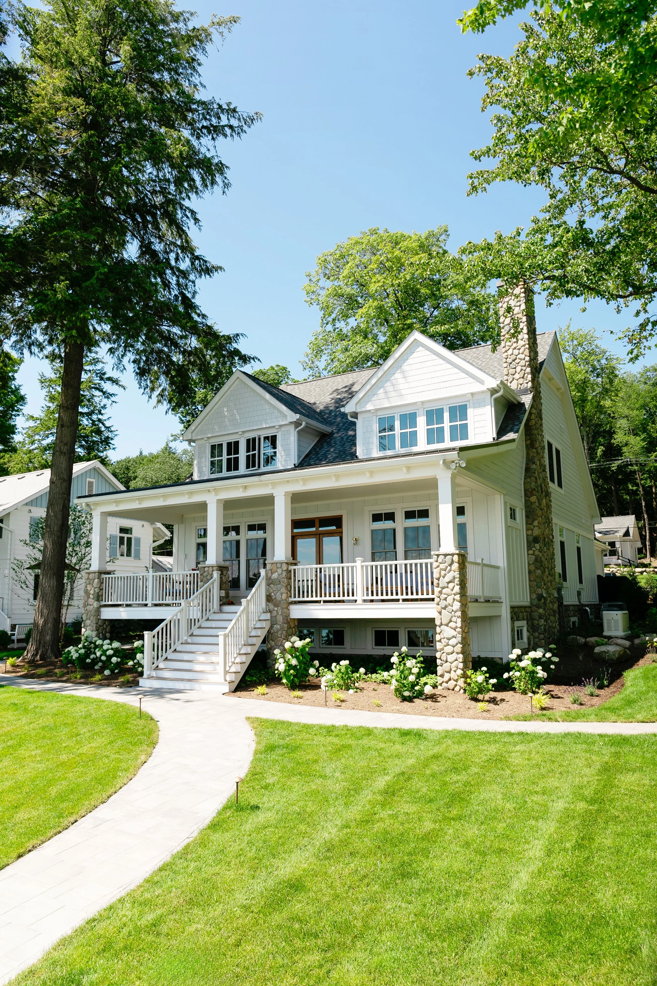 A white two-story house with a front porch and stairs, surrounded by green grass, flowering bushes, and tall trees under a blue sky.