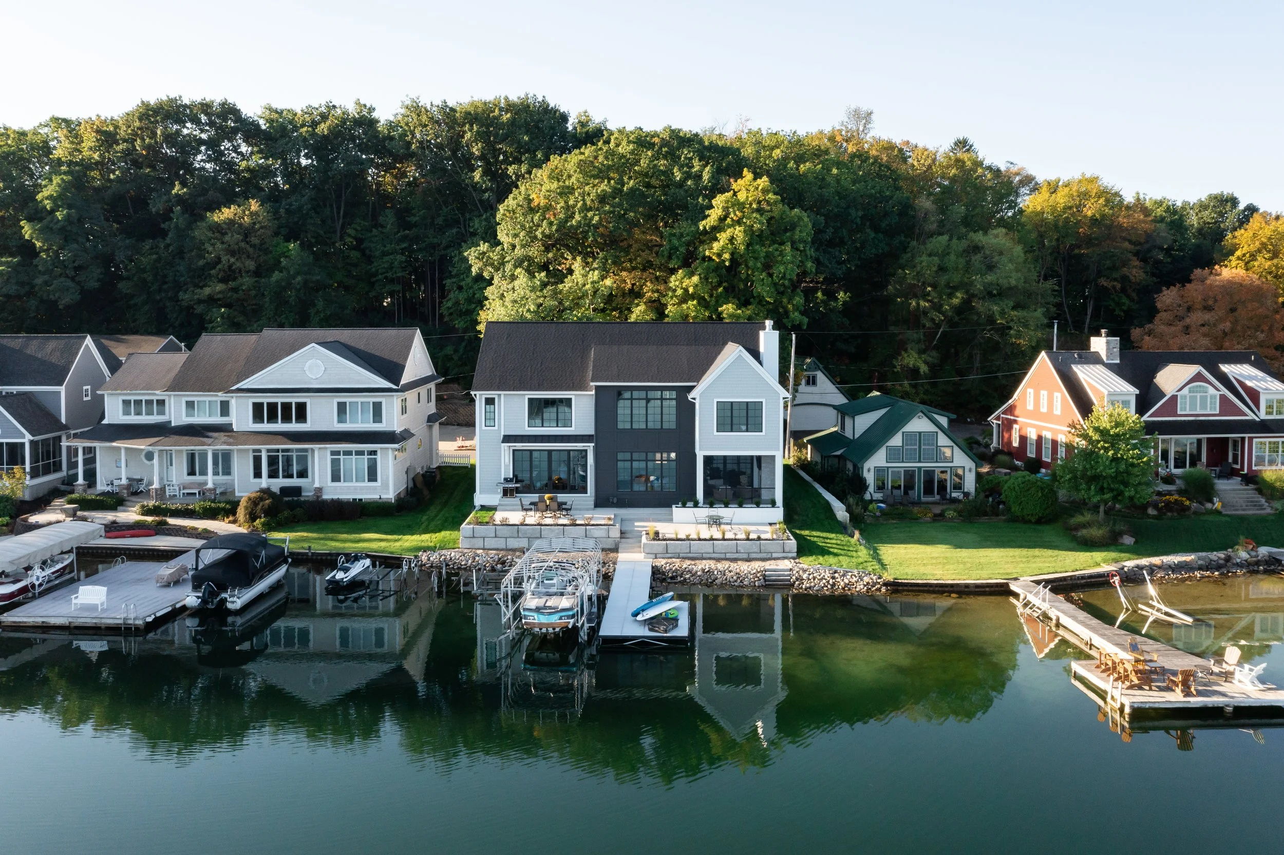 A row of modern houses with large windows and yards, situated along a calm waterway with docks and boats, surrounded by lush green trees in the background.
