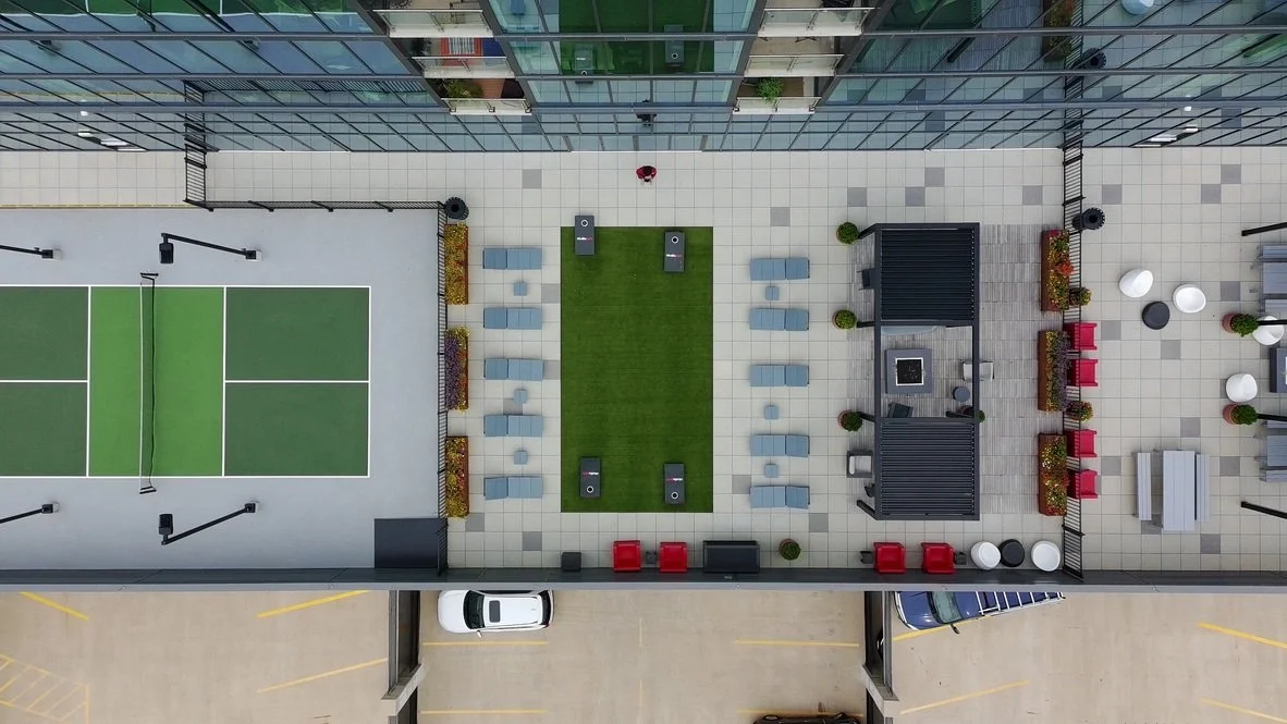 A rooftop sports and lounge area with a tennis court, seating, and landscaping, enclosed by glass buildings.