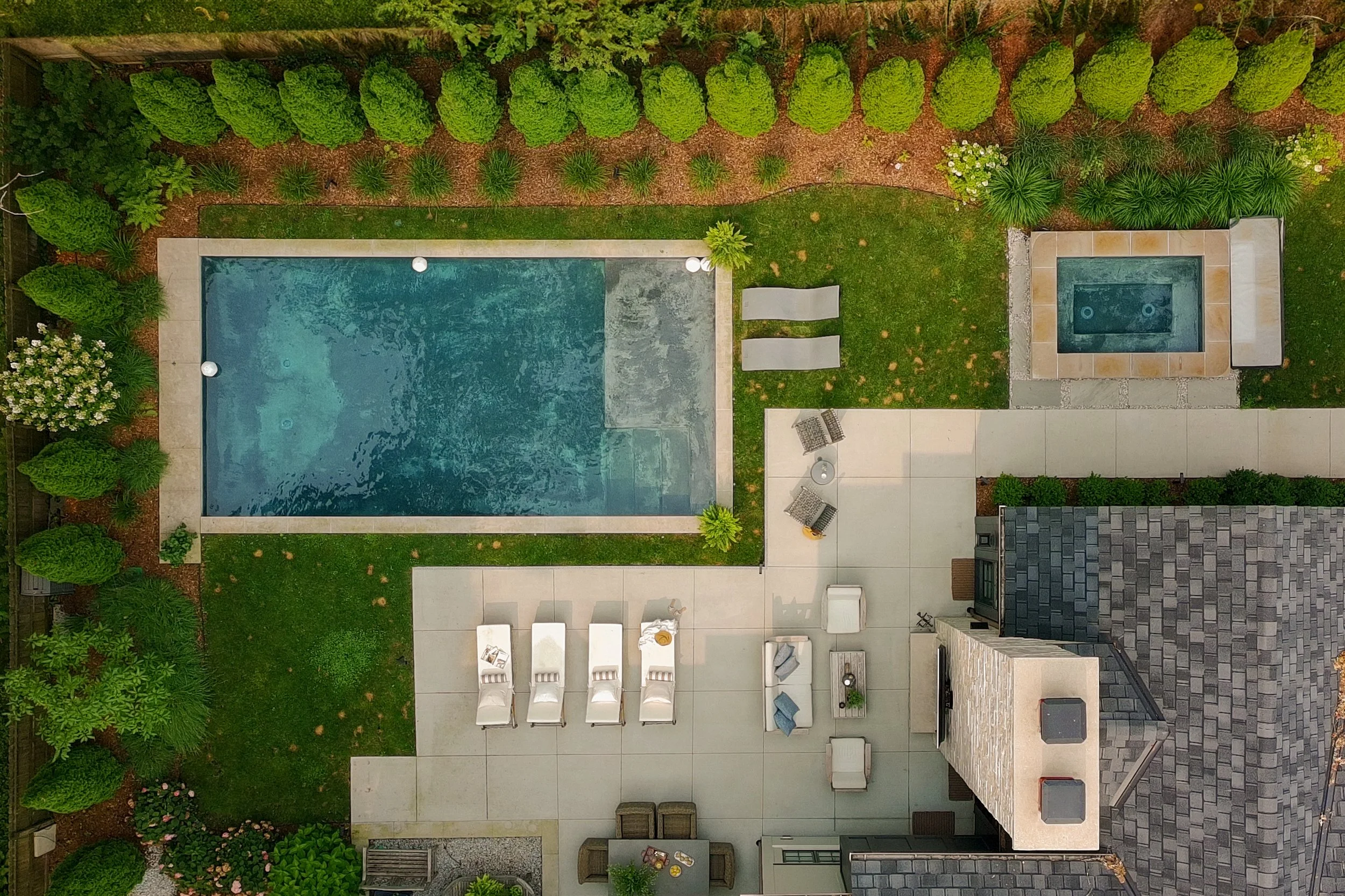 A backyard with a swimming pool, a hot tub, a patio with outdoor furniture, green grass, trees, and shrubs.