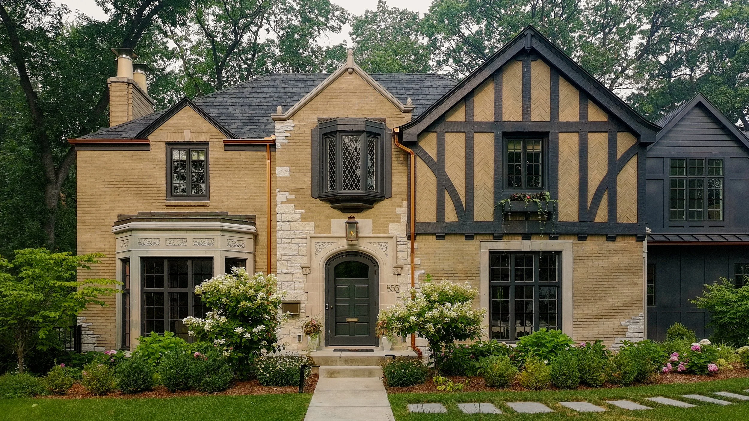 Front view of a large house with light brick and dark wooden accents, with a well-maintained garden and pathway leading to the front door.
