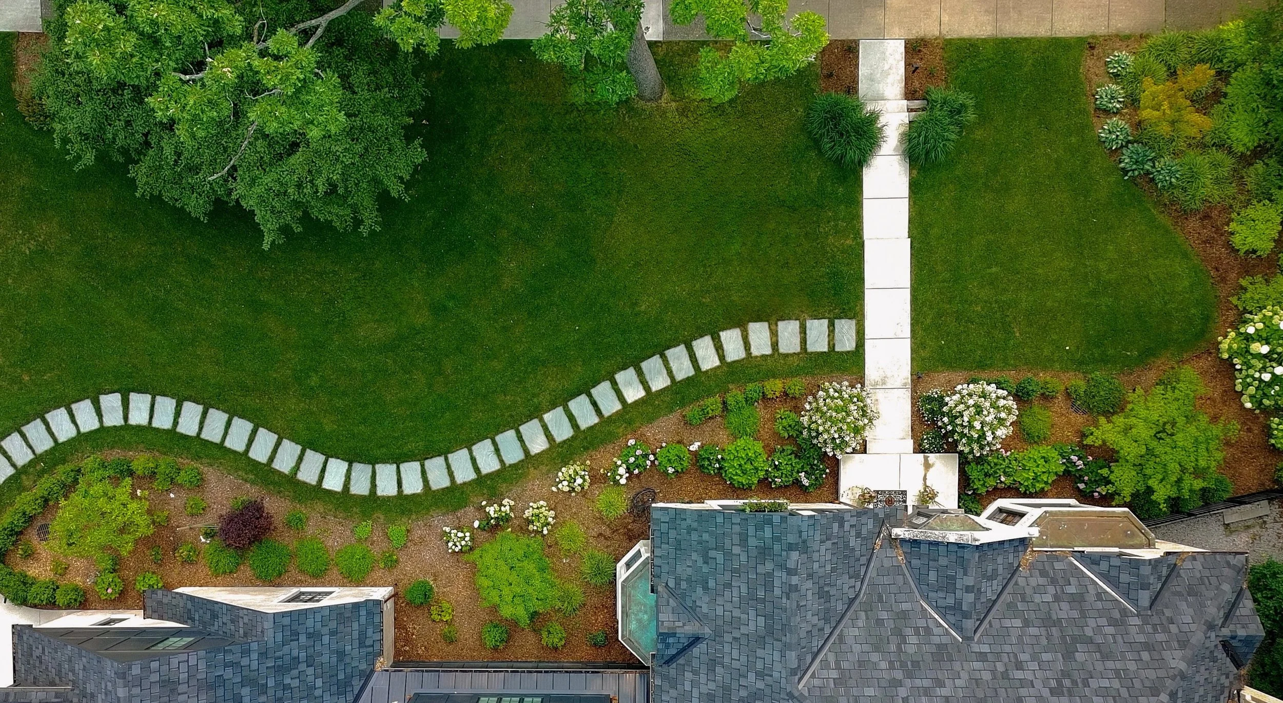 An aerial view of a backyard with a curved stone pathway and well-maintained green lawn, bordered by a variety of shrubs and trees, with parts of the house's roof visible at the bottom of the image.