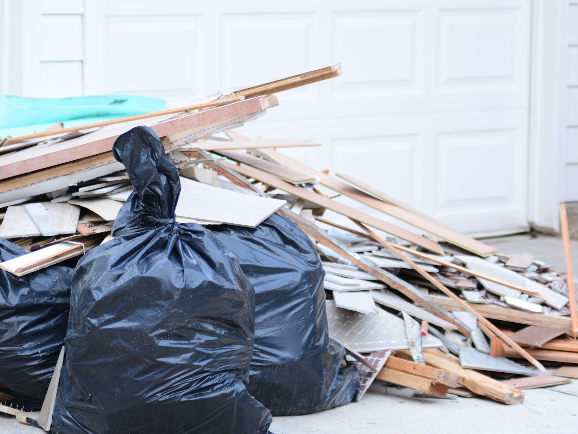 Pile of construction debris including wood and drywall next to black trash bags in front of a white garage door.
